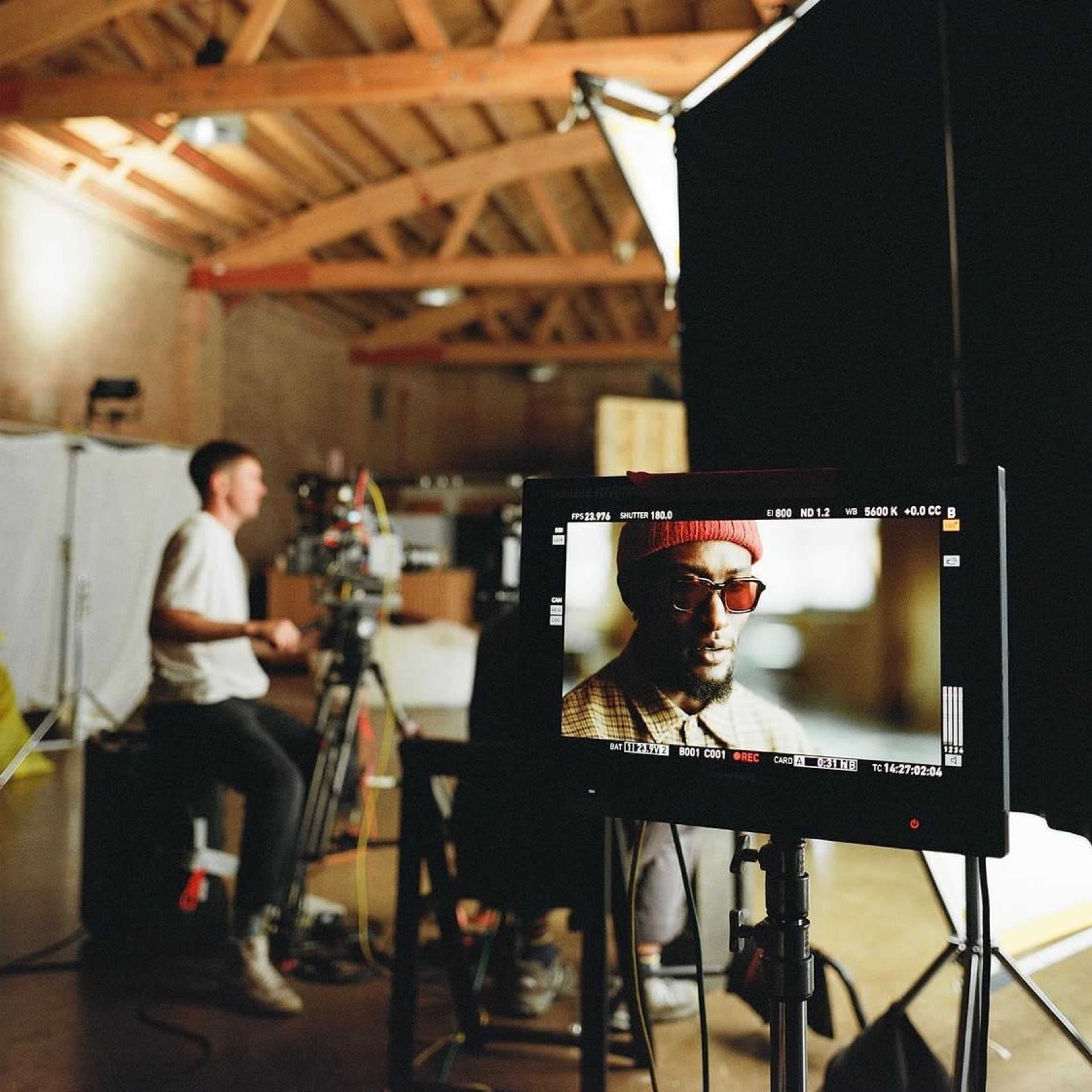 A film crew works in a studio, with one person operating a camera and another being filmed in front of a monitor.