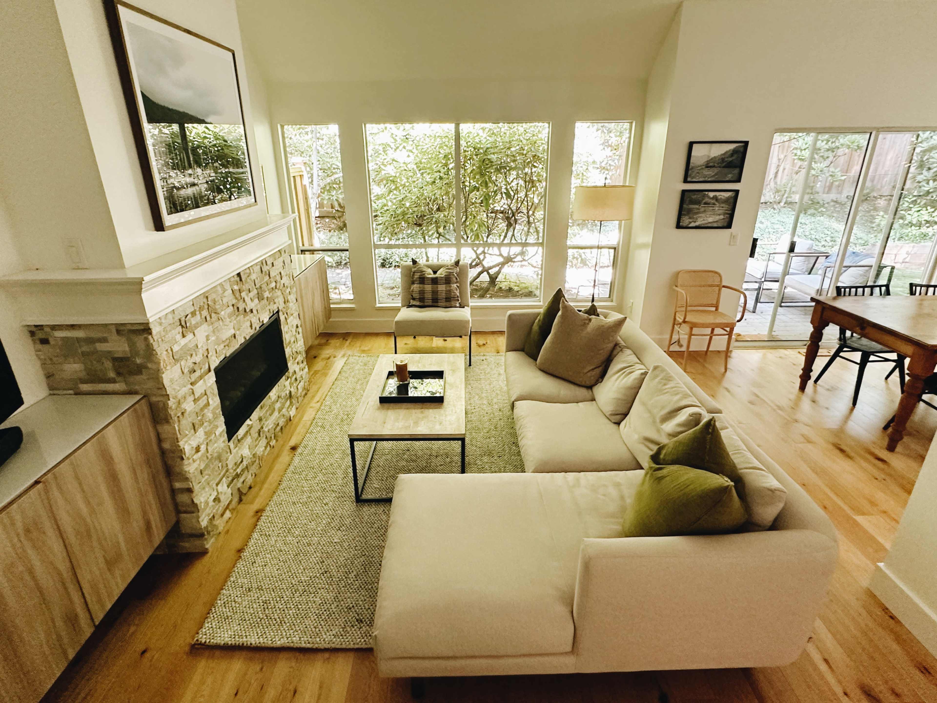 The image shows a modern living room with a sectional sofa, a coffee table, a stone fireplace, and large windows overlooking a garden.