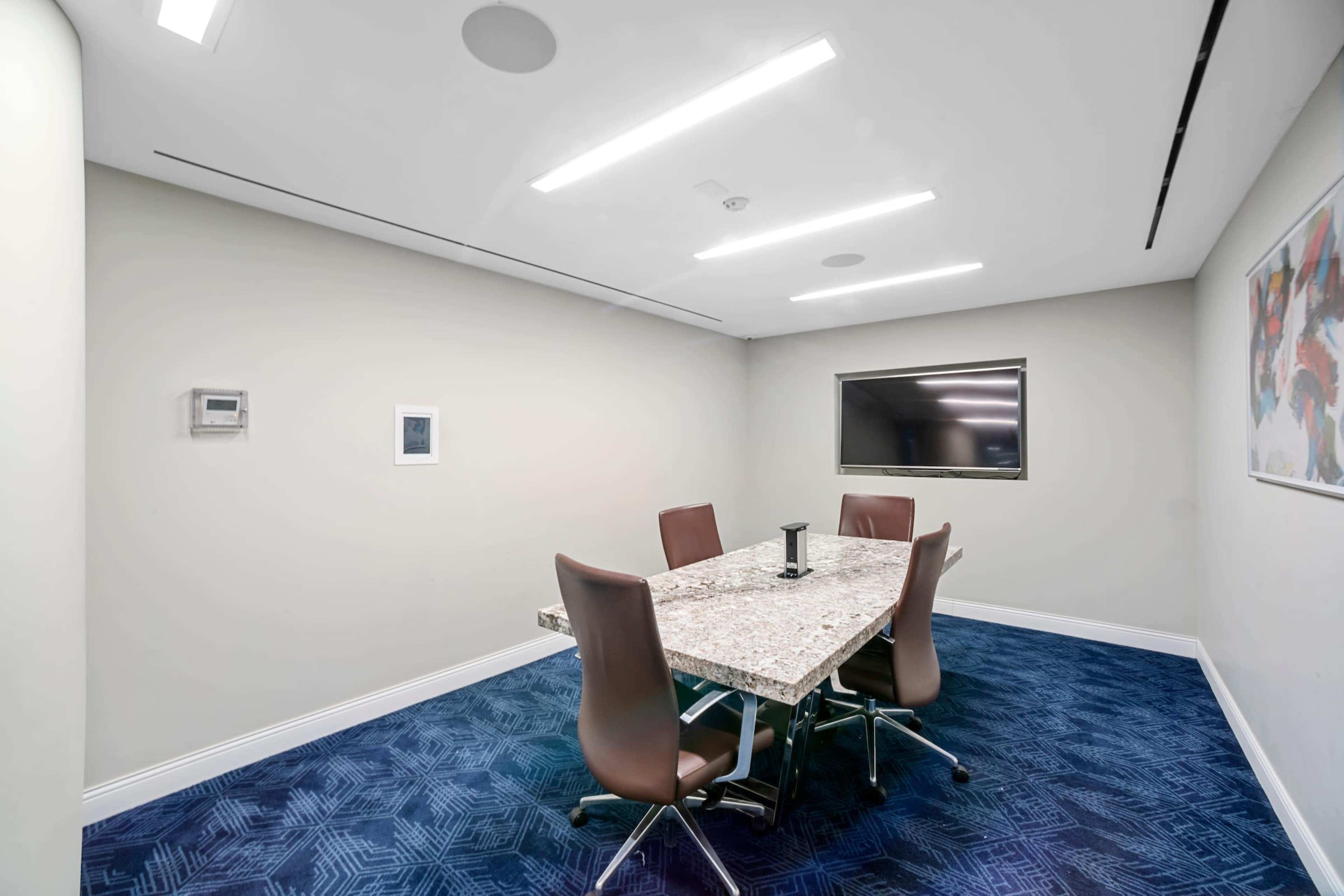 A conference room features a rectangular marble table surrounded by four brown chairs, with a large screen mounted on the wall and blue carpet covering the floor.