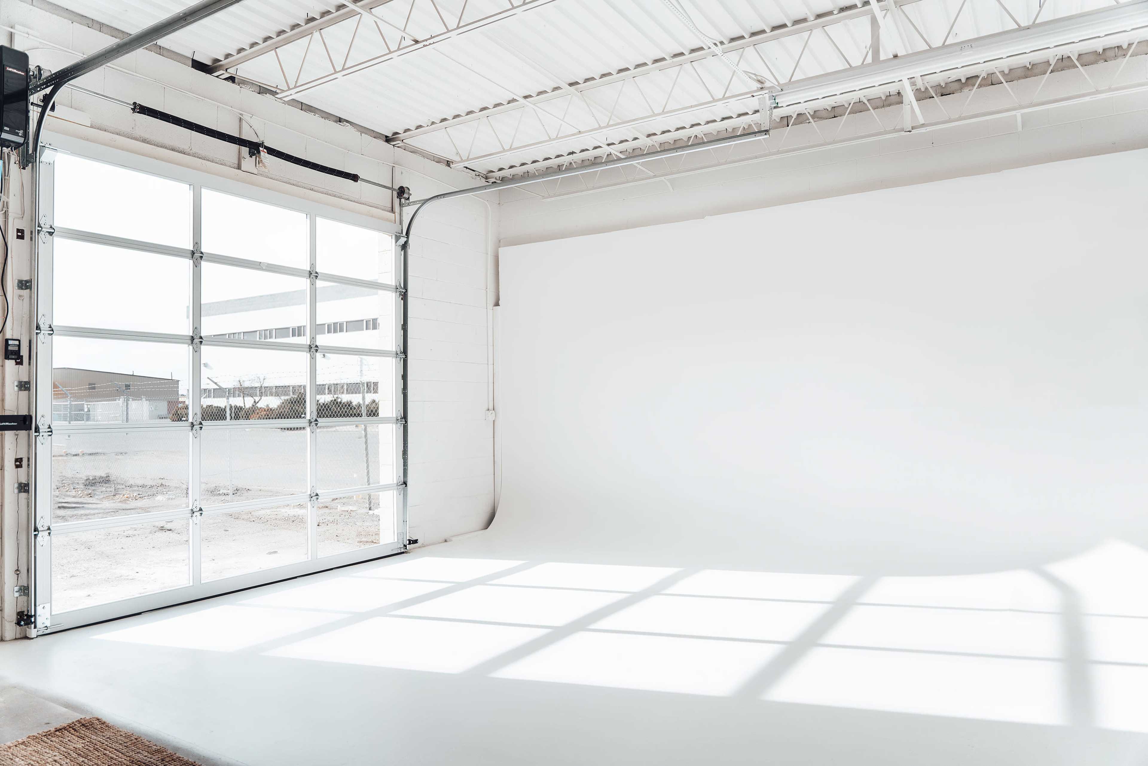 A spacious studio with a large garage door open to a well-lit interior, featuring a plain white backdrop and sunlight casting shadows on the floor.