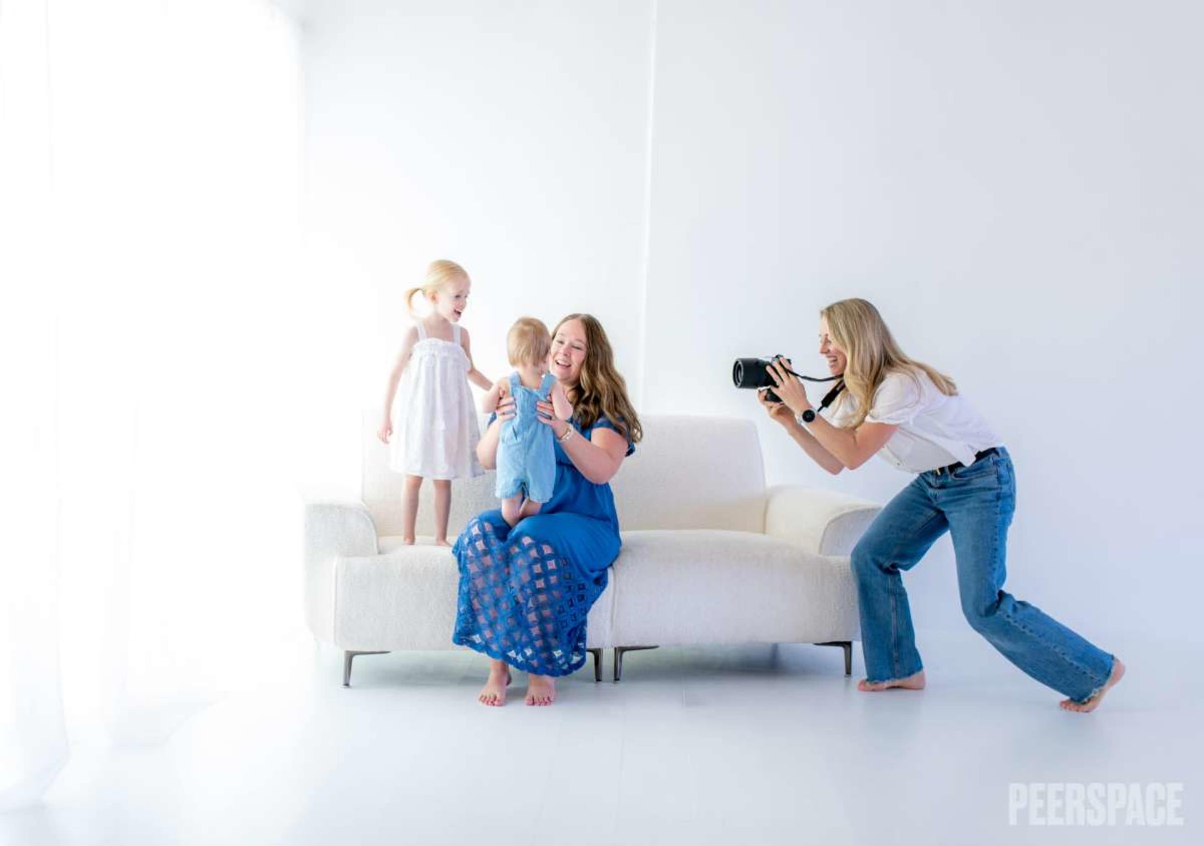 A woman holds a baby on a couch while another woman photographs them, and a young girl stands beside them.