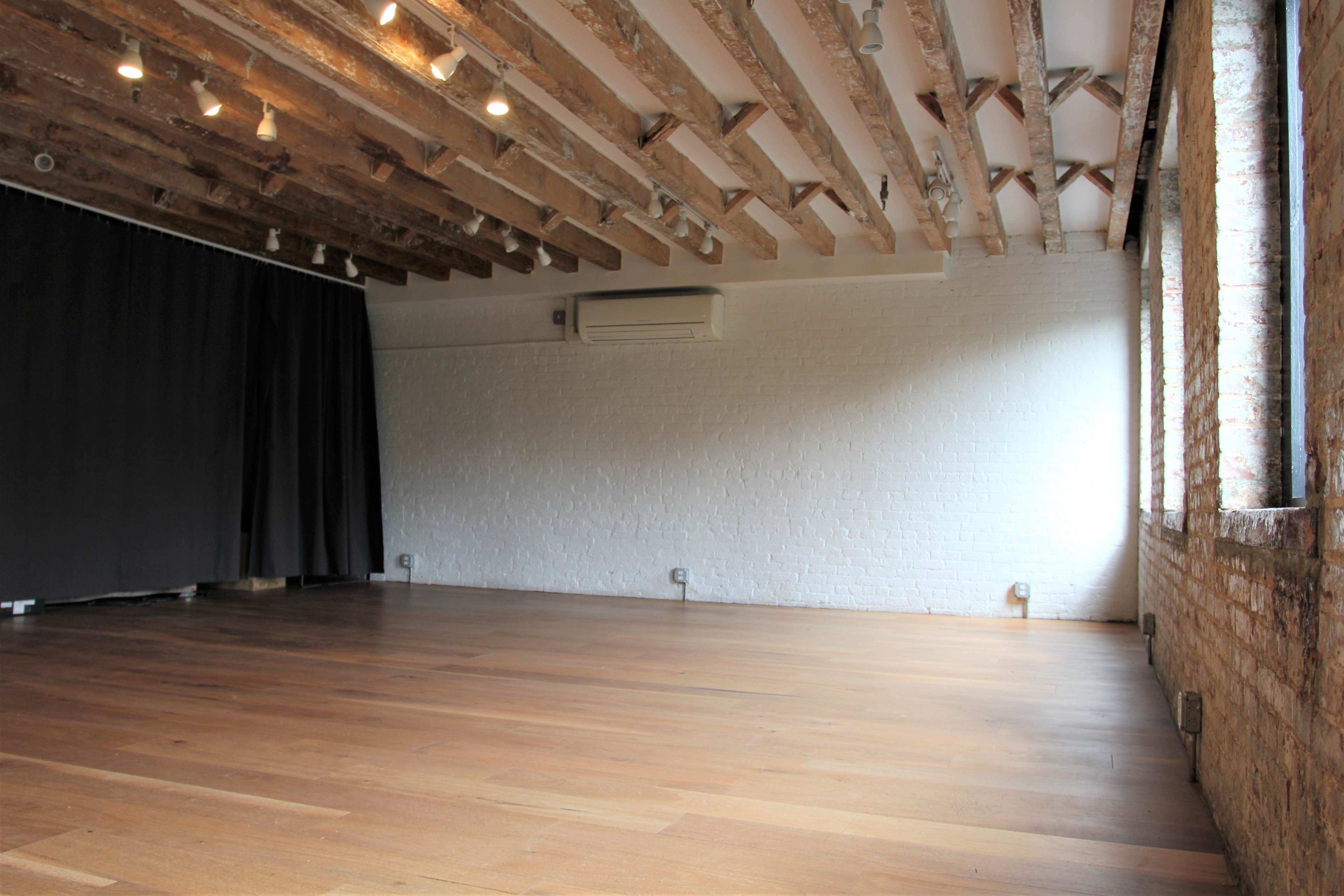A spacious interior room with exposed wooden beams, hardwood flooring, and large windows illuminating a white brick wall.