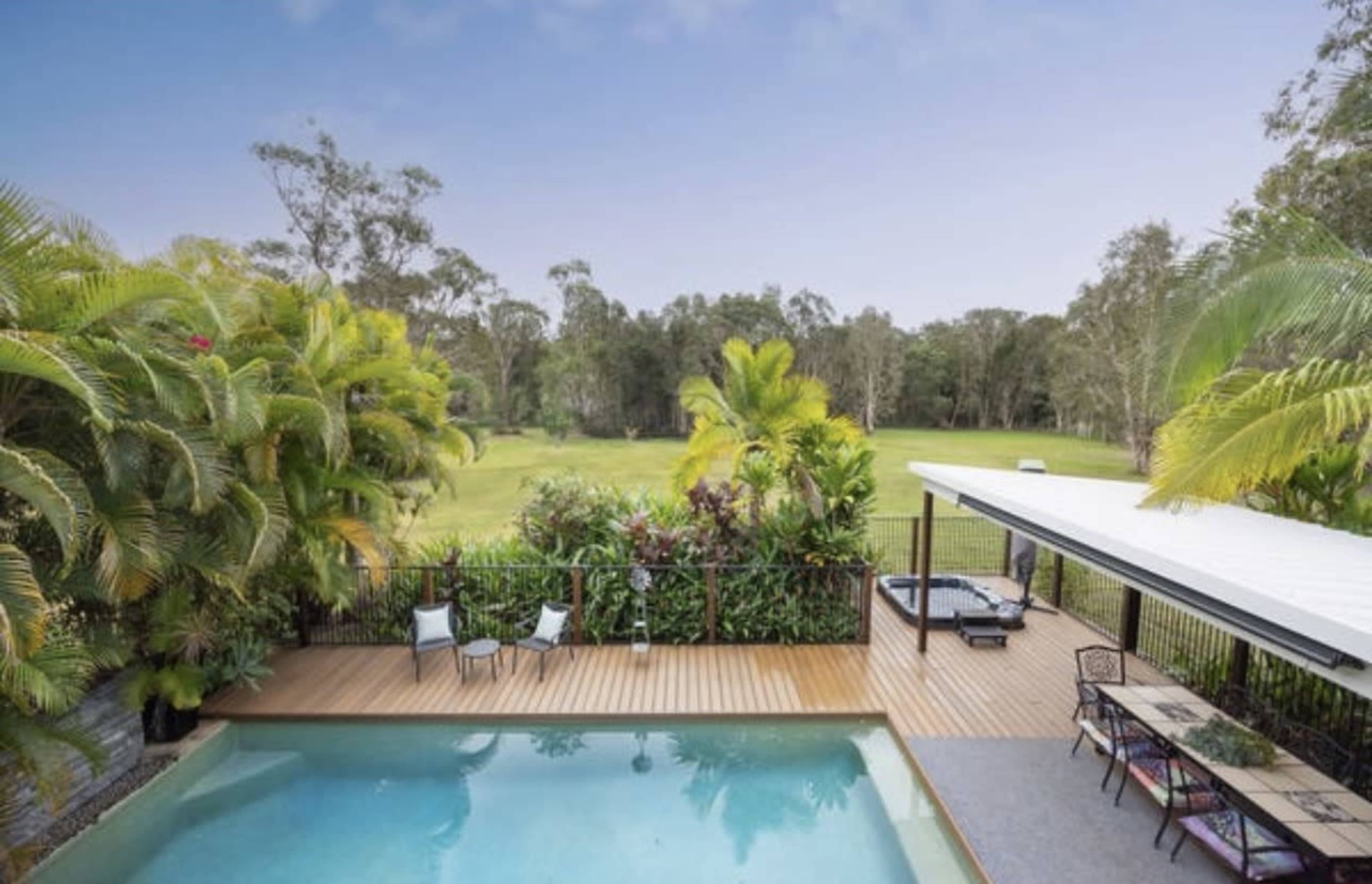 A swimming pool surrounded by tropical plants and a wooden deck features seating areas and overlooks a lush green landscape.