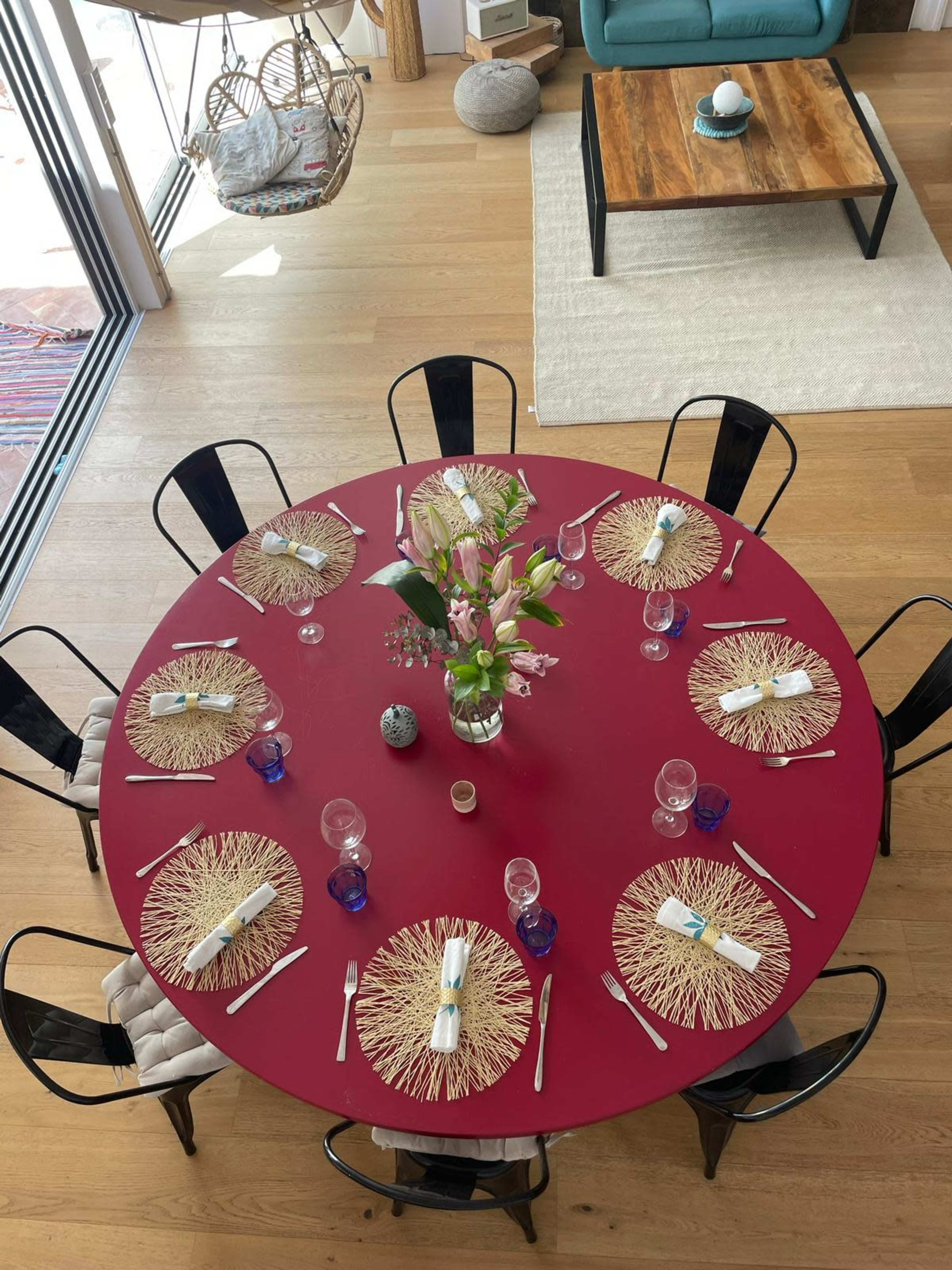 A round dining table with a red surface is set for a meal, featuring floral centerpieces, glassware, and woven placemats.