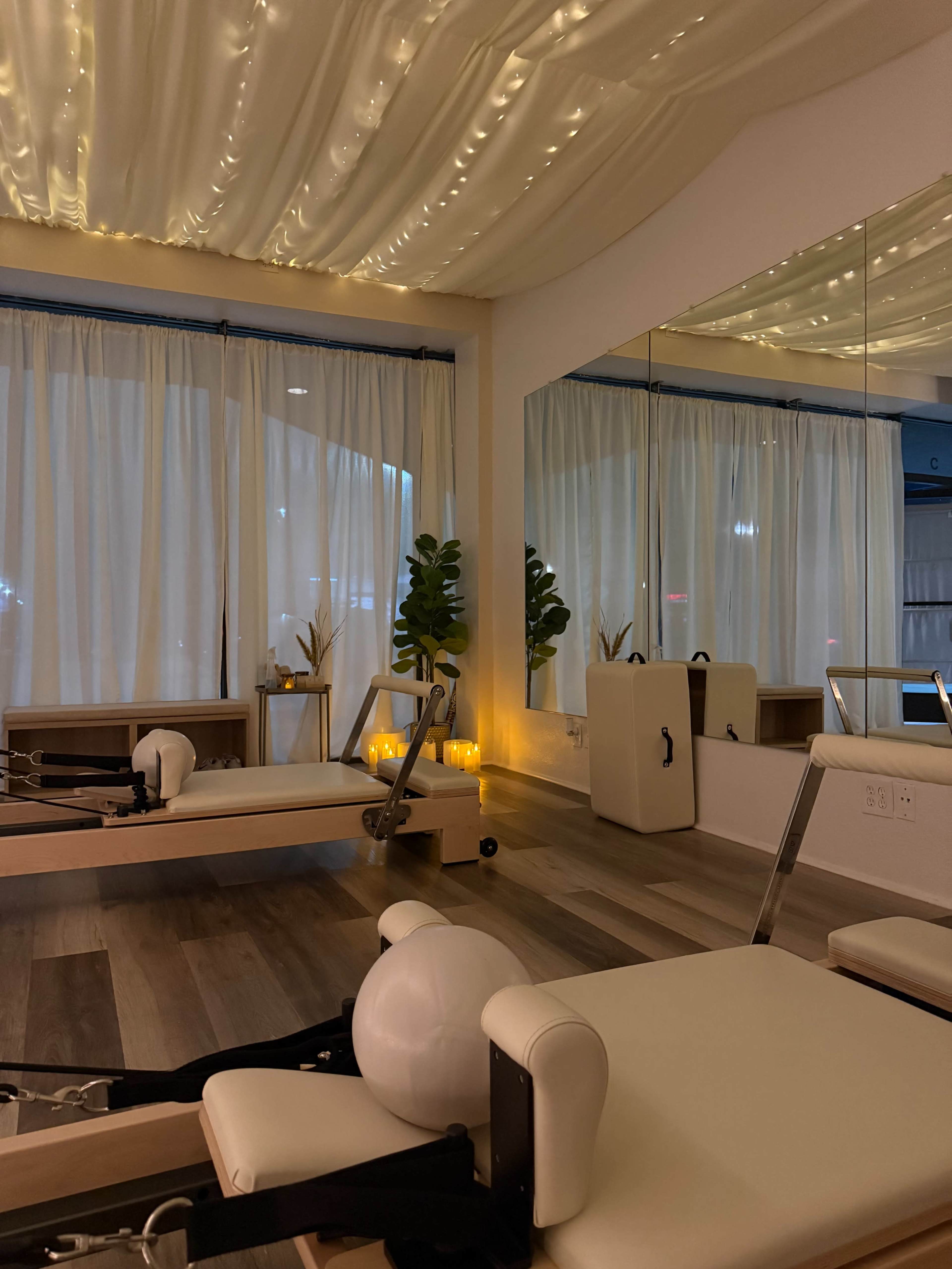 The image shows a serene Pilates studio with low lighting, featuring reformer machines, soft curtains, and decorative plants.