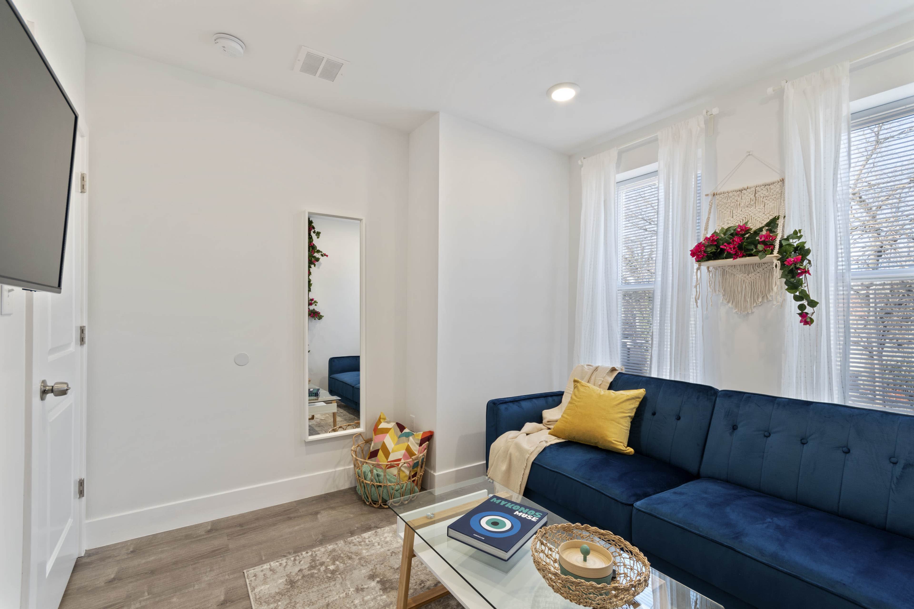 A modern living room features a blue sofa, a glass coffee table, a large mirror, and decorative plants by the windows.
