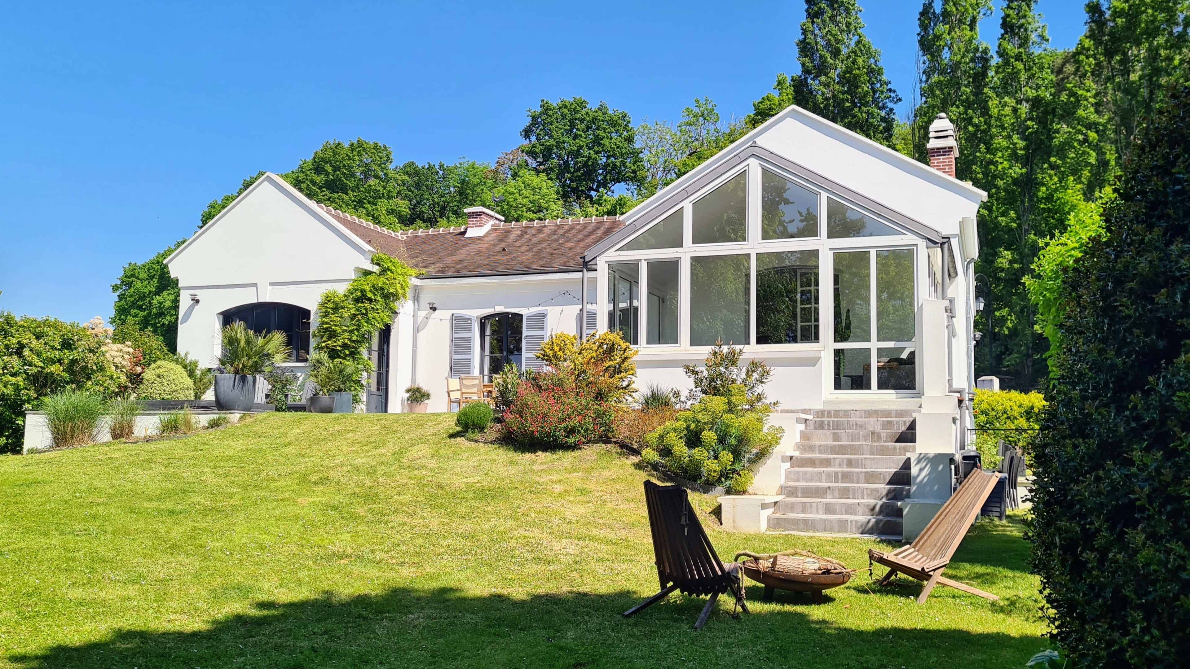 A modern house with a large glass-enclosed patio sits on a green hillside surrounded by trees and landscaped gardens.