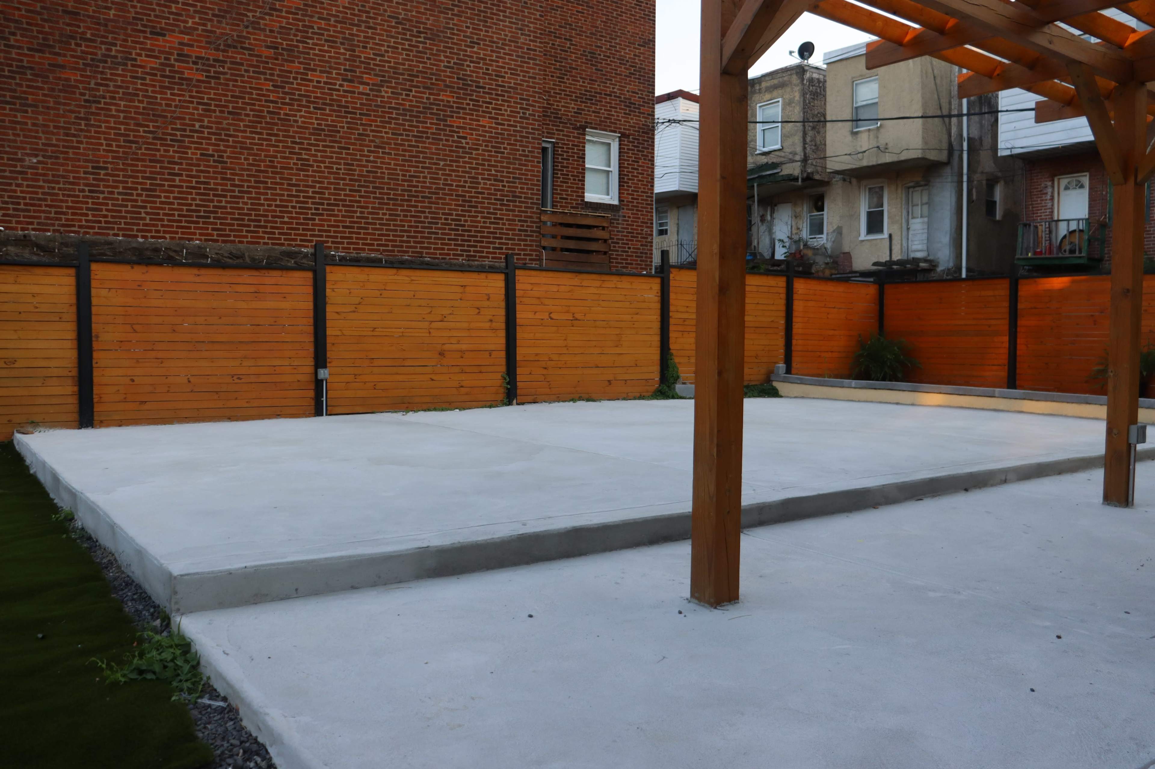 The image shows a newly prepared concrete slab in a backyard, surrounded by wooden fencing and a pergola.