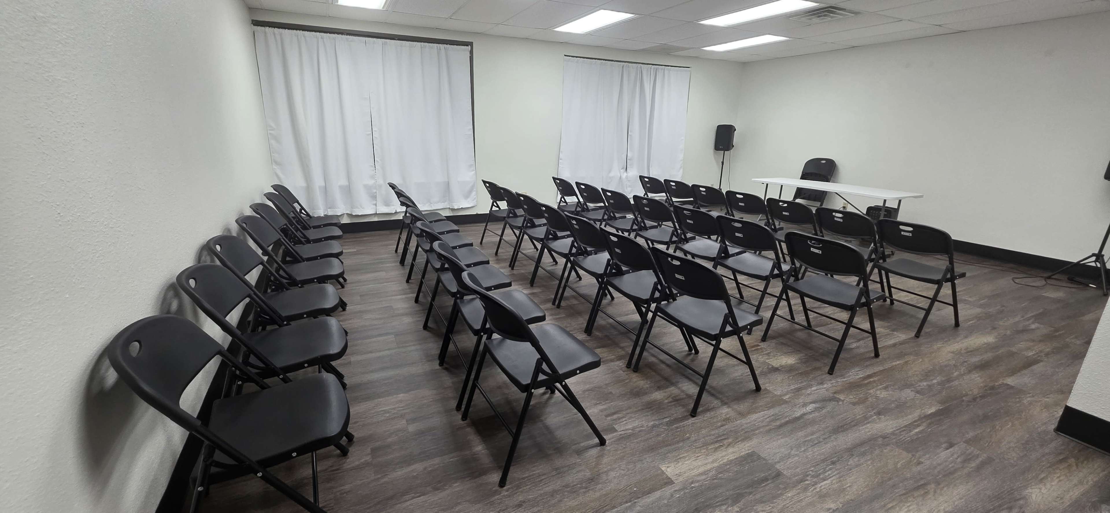 The image shows a spacious room arranged with rows of black folding chairs facing a table, with white curtains covering the windows.