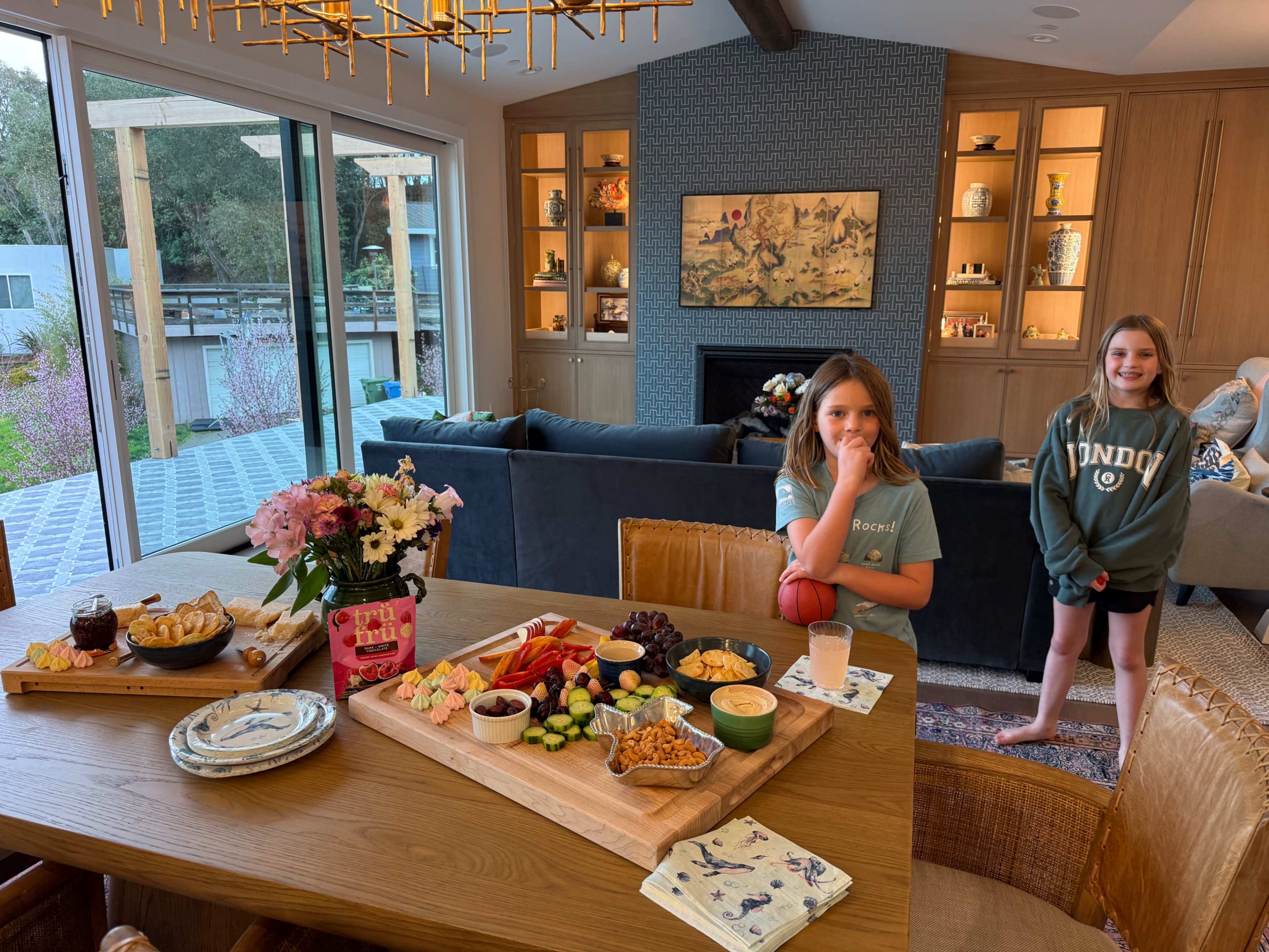A dining table is set with a variety of snacks and drinks, while two girls stand nearby in a well-decorated living room with large windows and modern furnishings.