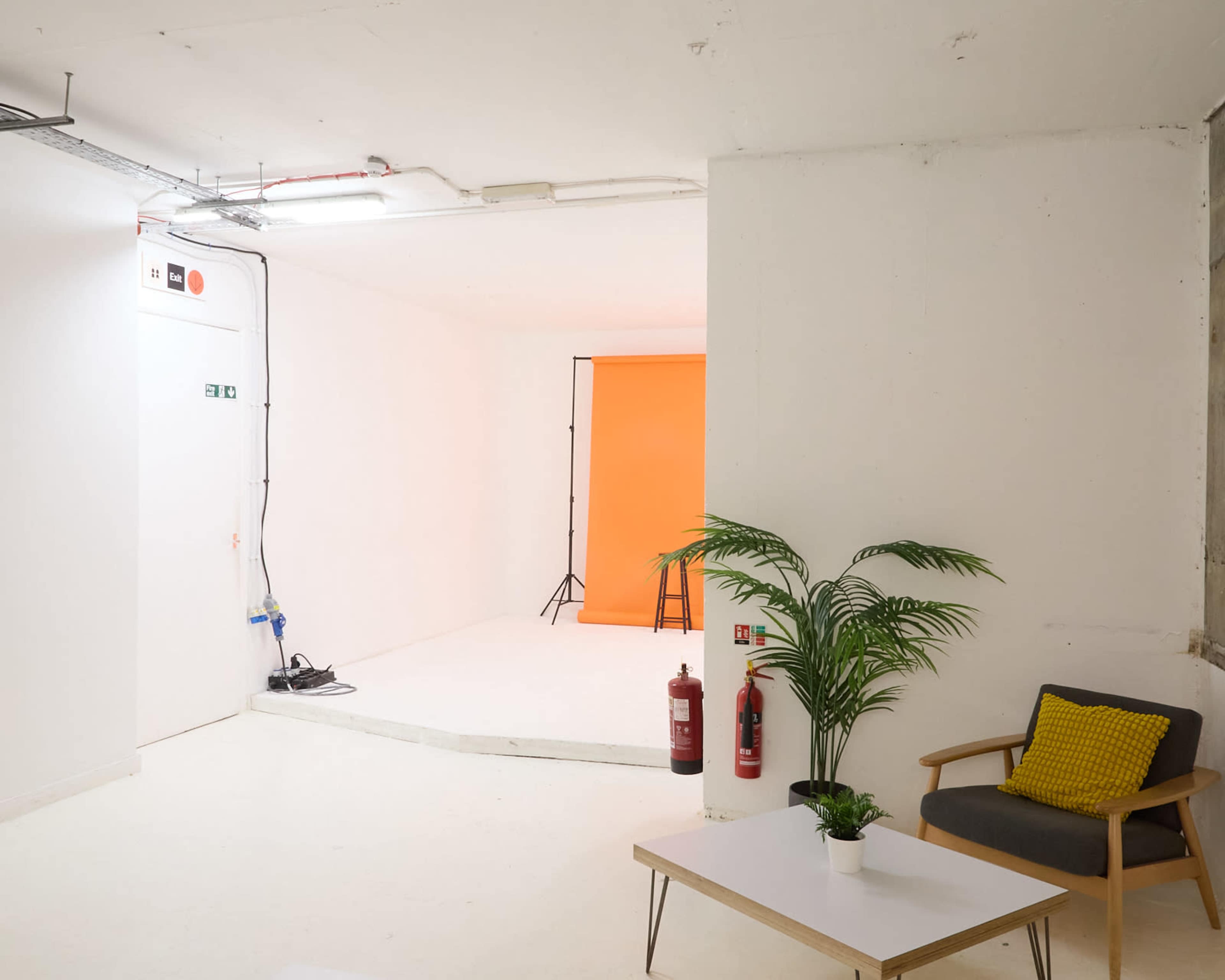 The image shows a minimalist studio space with a white wall, a raised platform, a chair, and a coffee table, along with a potted plant and fire extinguishers.