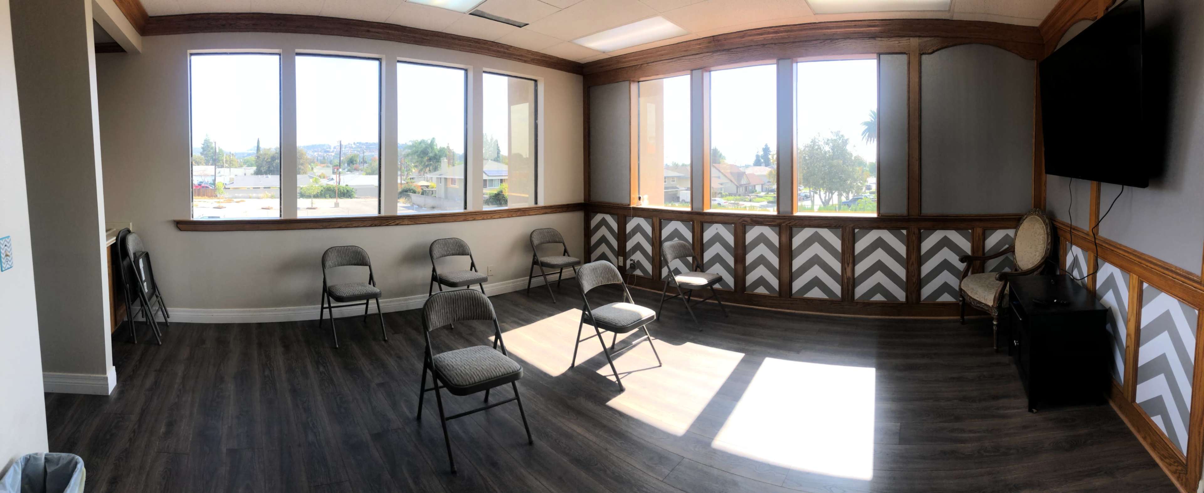 A spacious room with several folding chairs arranged on a wood-style floor, large windows providing natural light, and a television mounted on the wall.