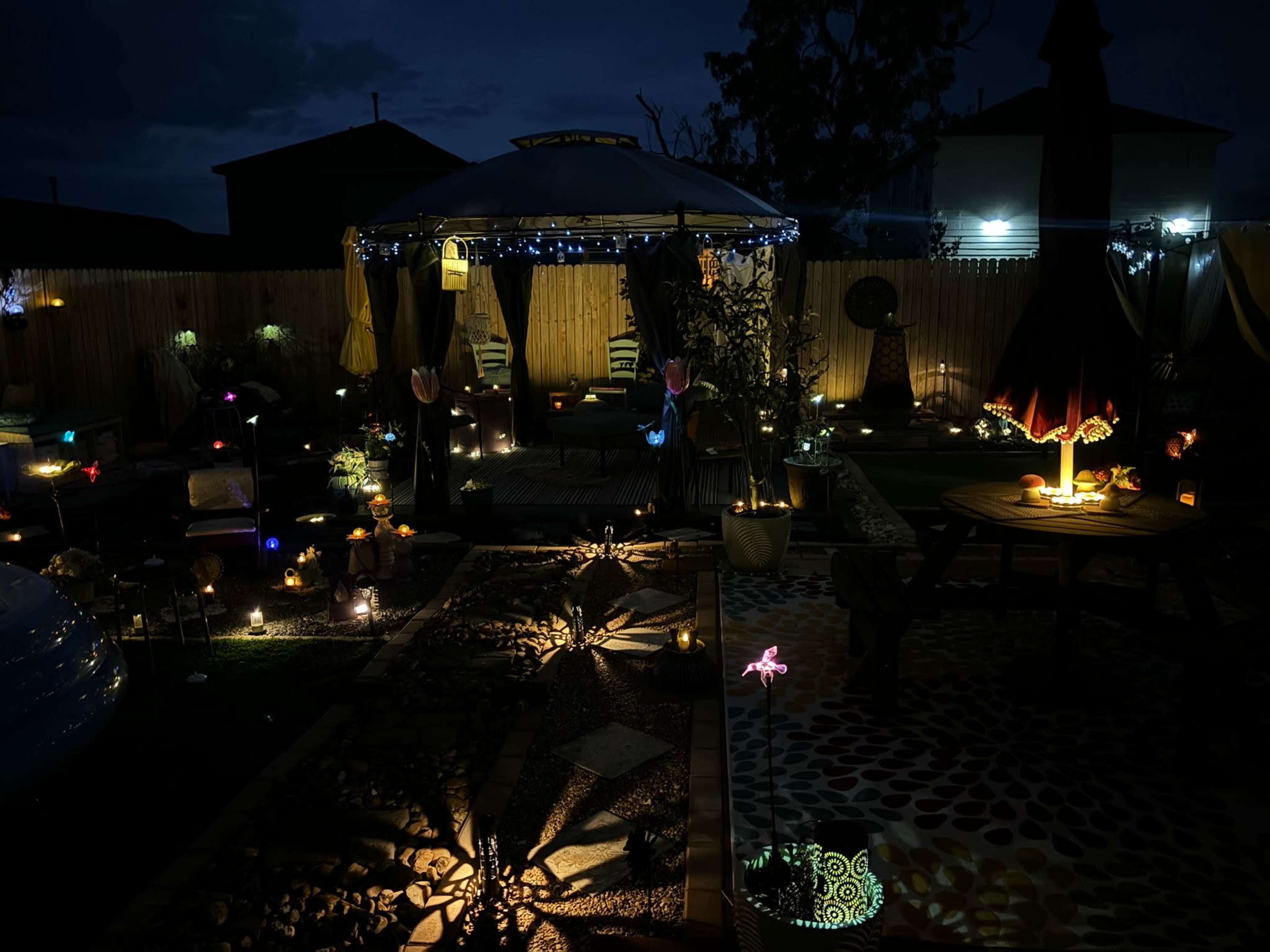 The image shows a backyard illuminated by various decorative lights and lanterns, featuring a gazebo surrounded by plants and seating areas.