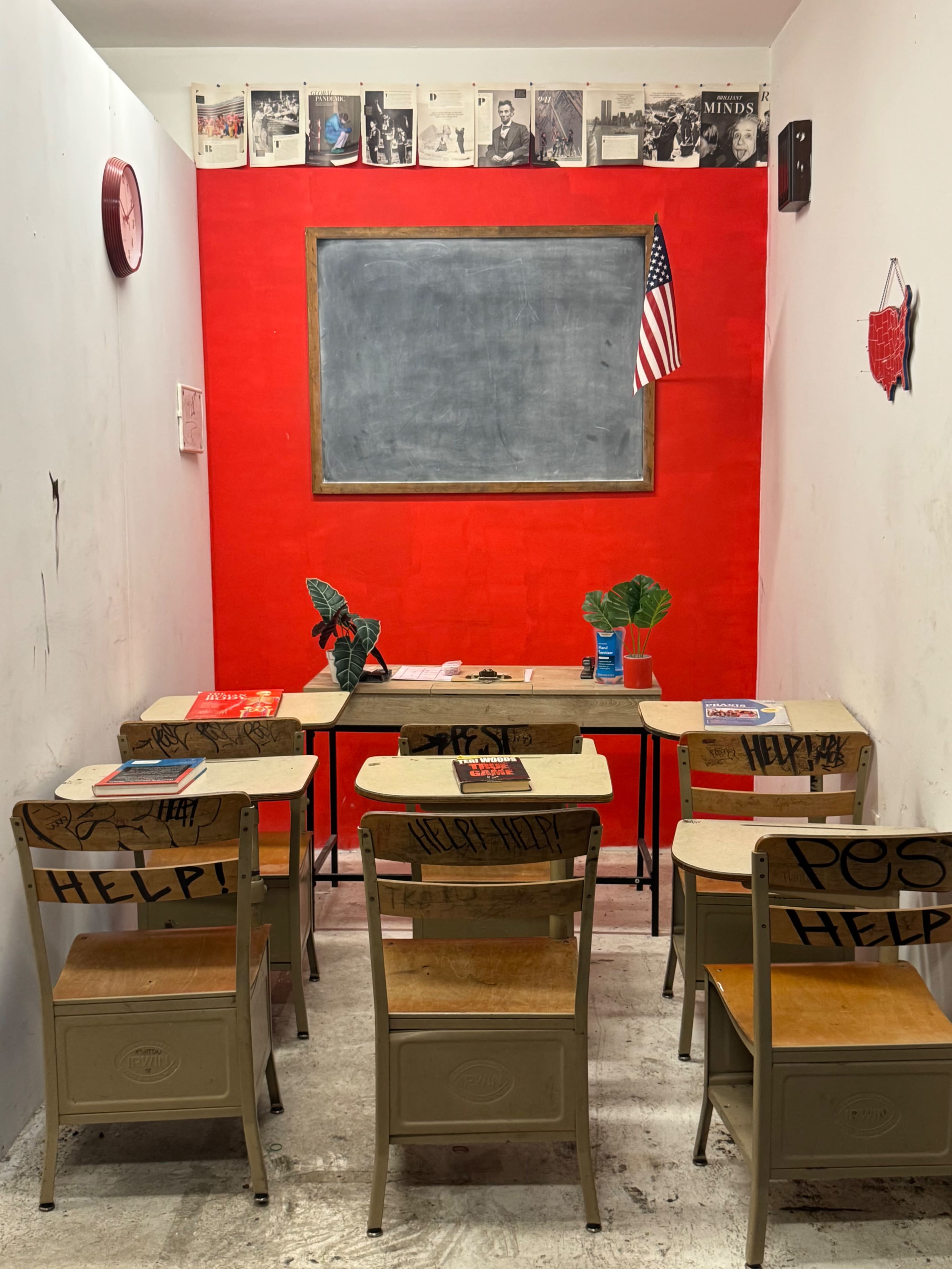 The image shows a small classroom with empty wooden desks and chairs facing a blank chalkboard, against a red wall adorned with an American flag and various photographs.