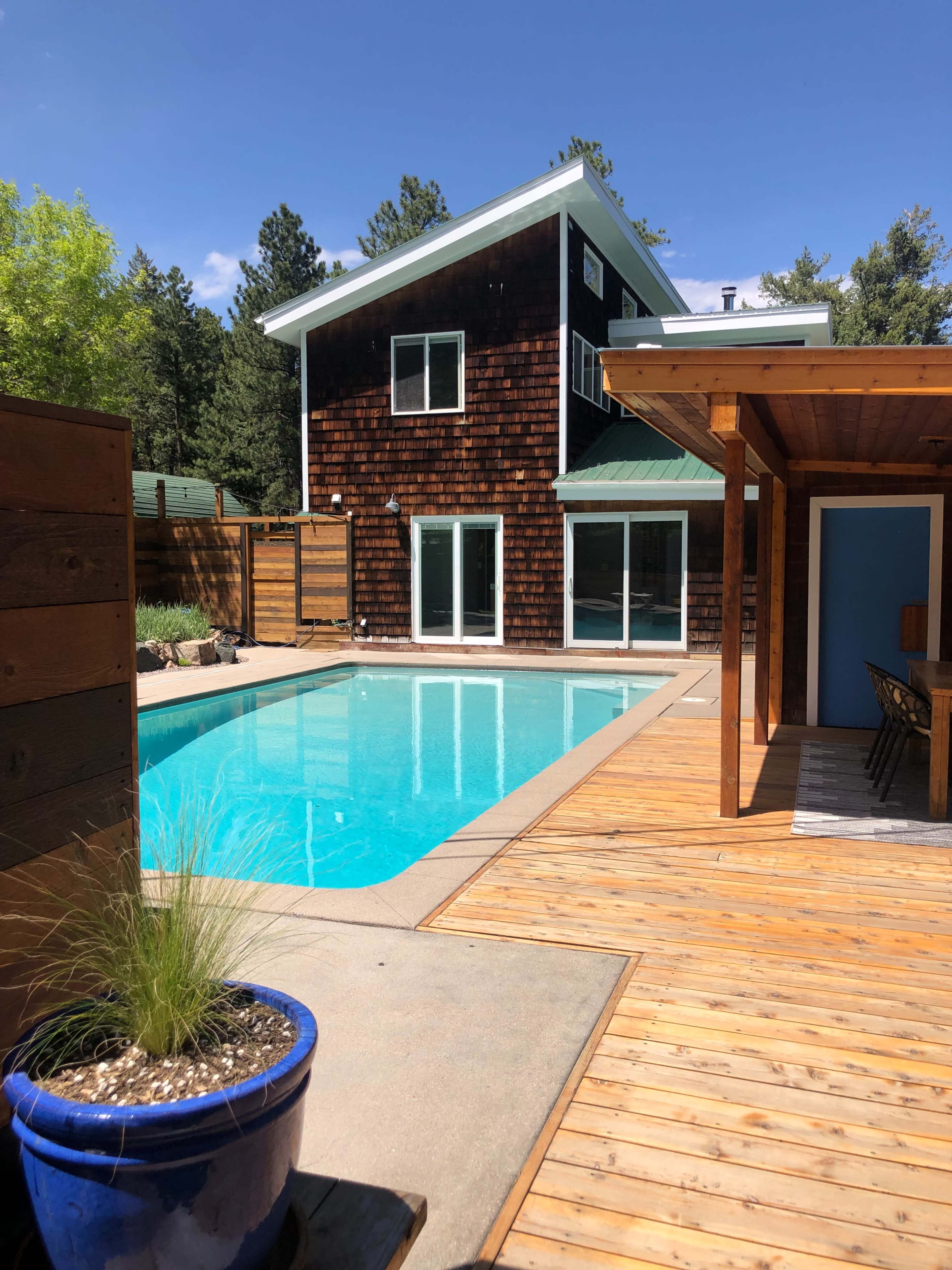 The image shows a modern house with a wooden exterior, a swimming pool in the foreground, and a deck area that includes a dining space.