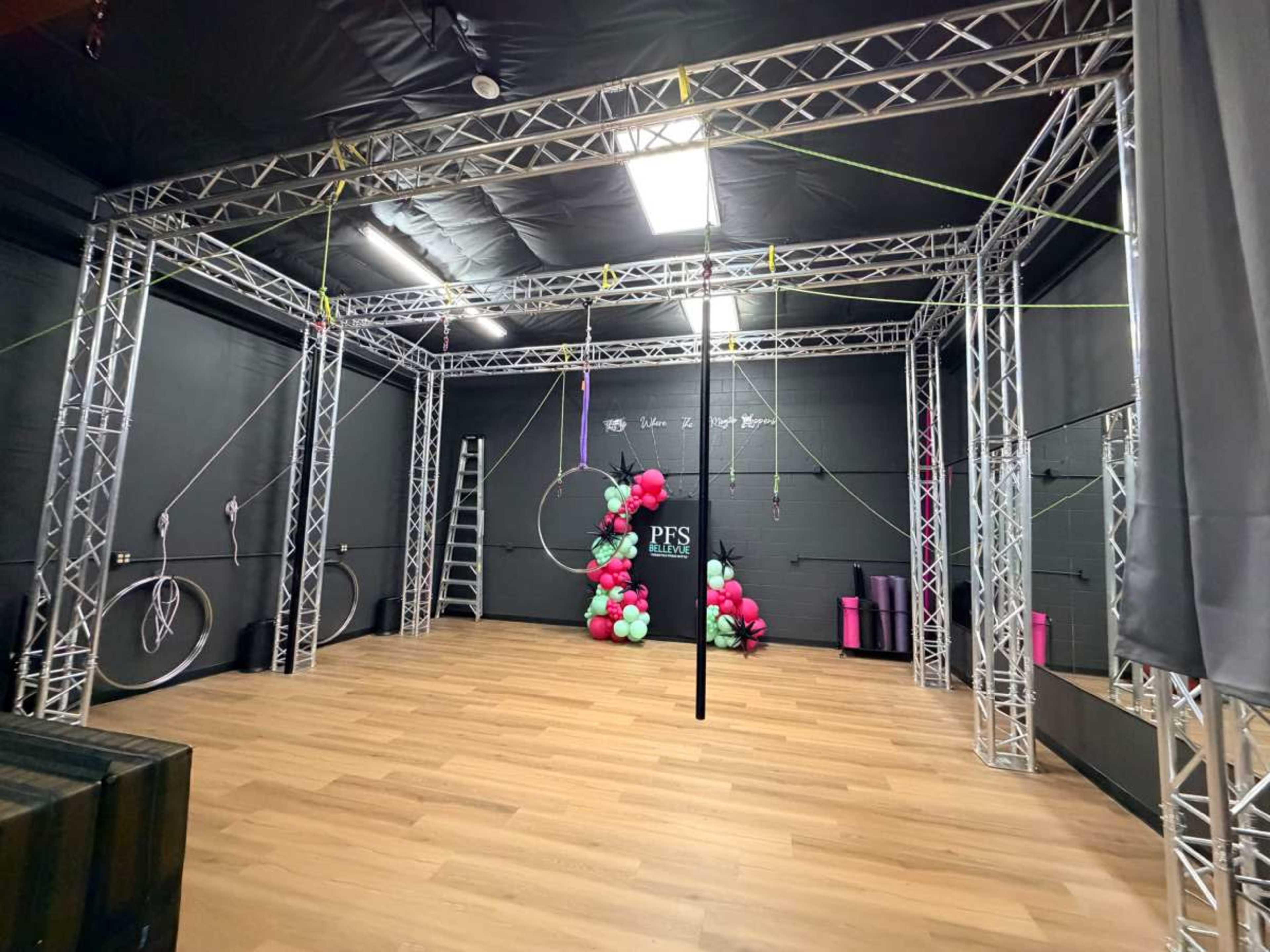 The image shows a spacious indoor fitness studio featuring metallic rigging structures, exercise equipment, and a decorative backdrop of balloons in pink and green.