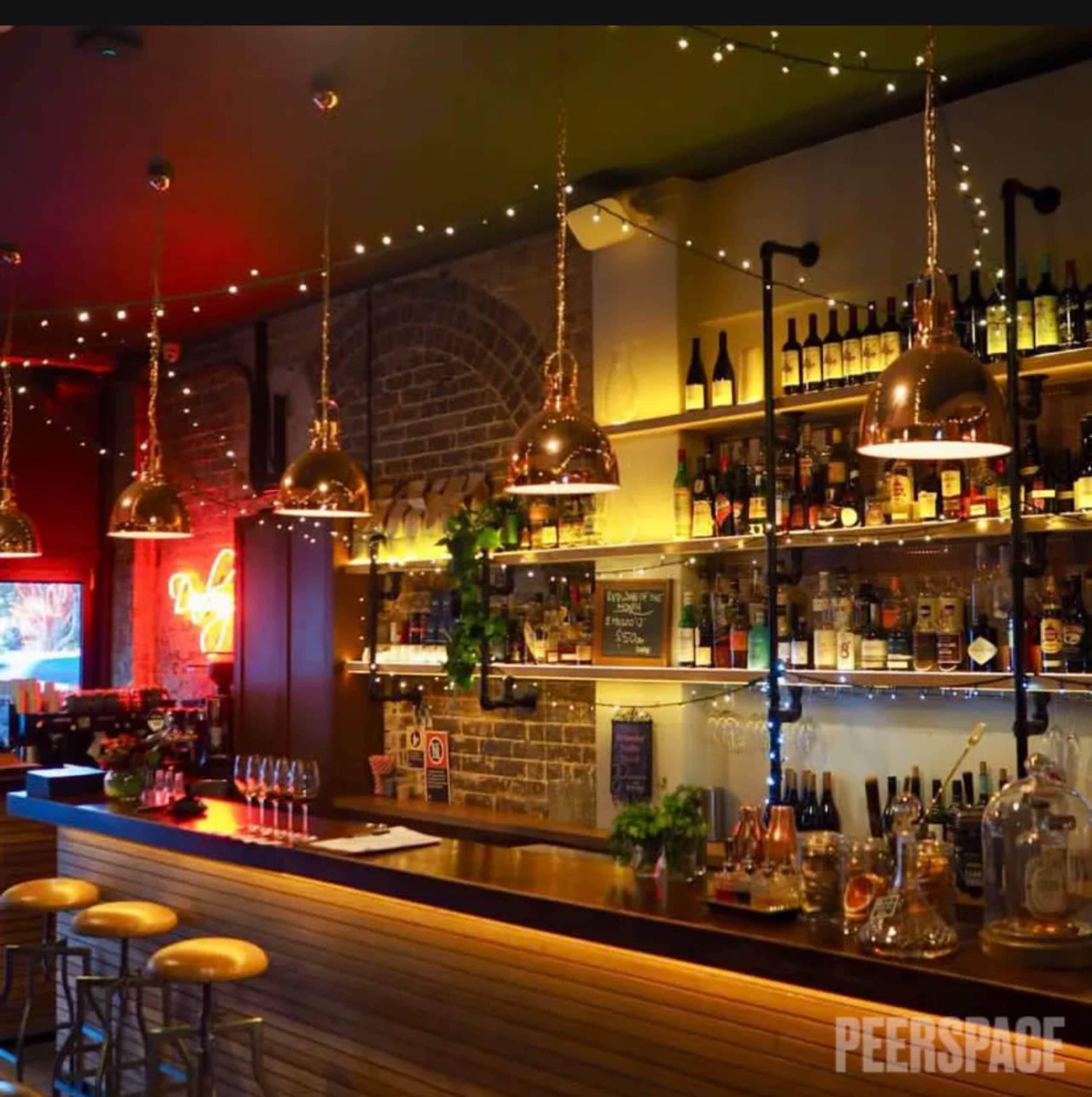 The image shows a dimly lit bar featuring a wooden counter, copper pendant lights, and shelves lined with various bottles.