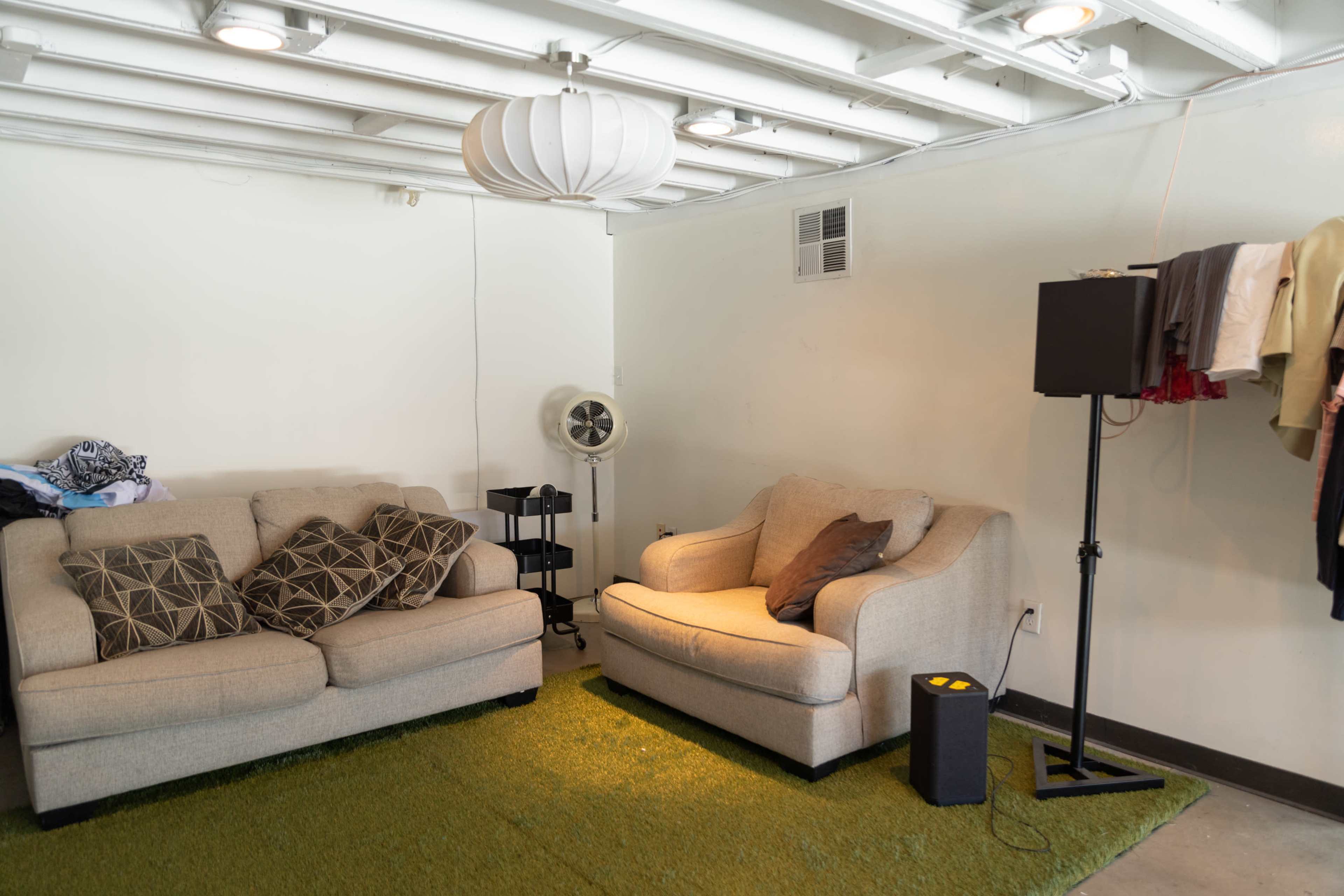 The image shows a neatly arranged living room with two sofas, a fan, a clothing rack, and a green rug on the floor.
