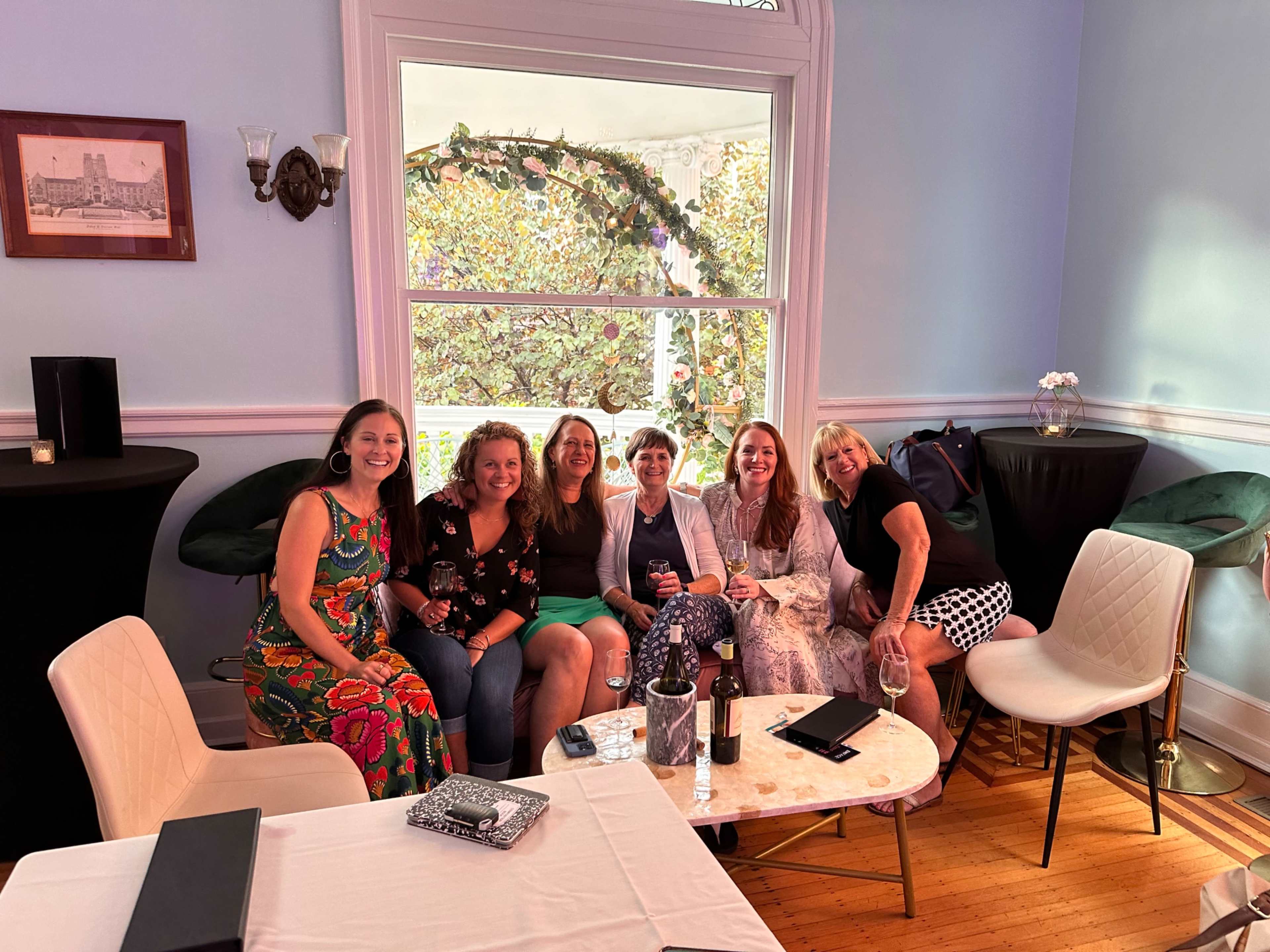 A group of six women is seated together in a brightly lit room with large windows and decorative greenery.