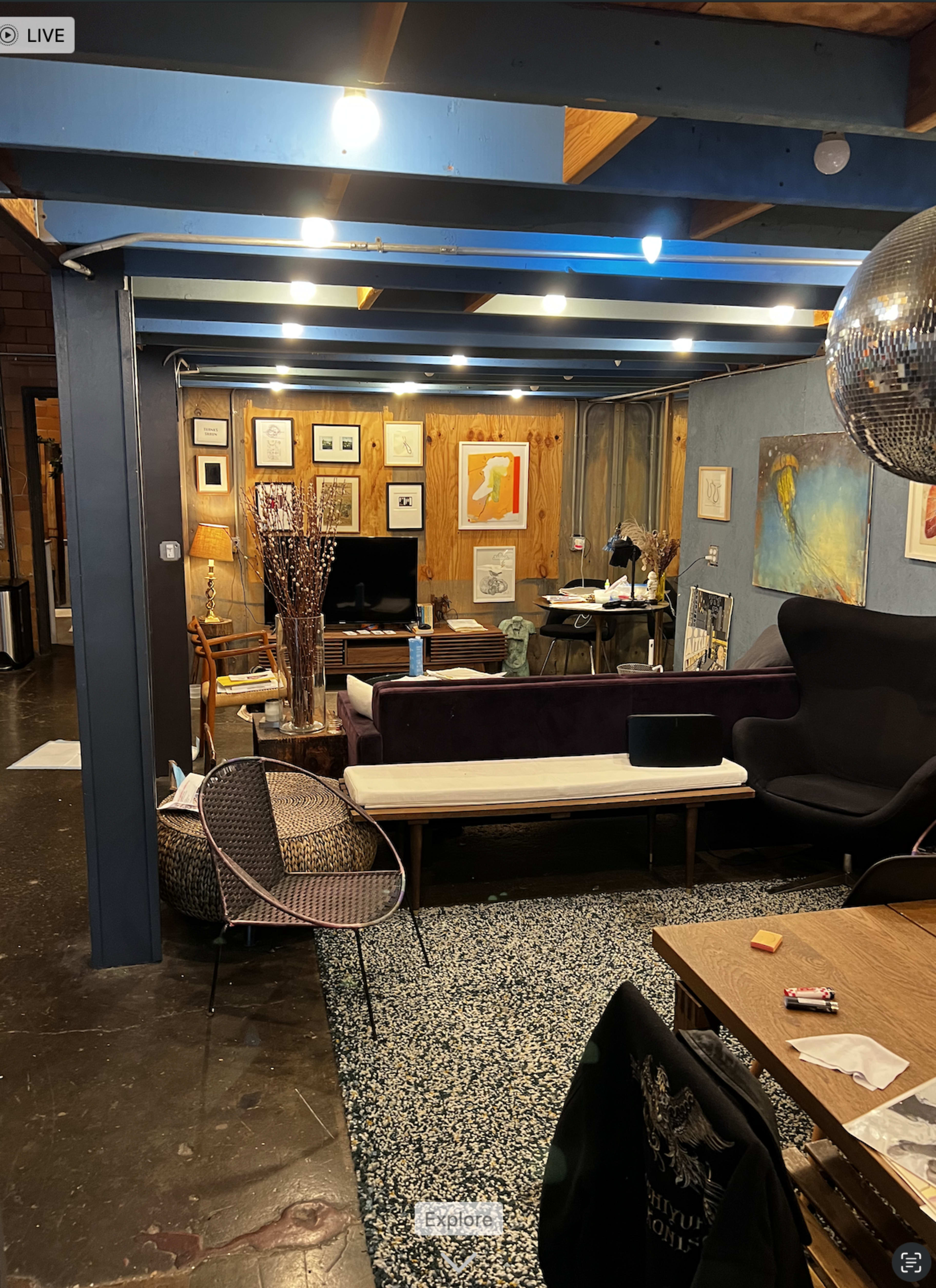 The image shows a cozy, eclectic living space with layered textures, framed artwork on the walls, and a mix of seating options.