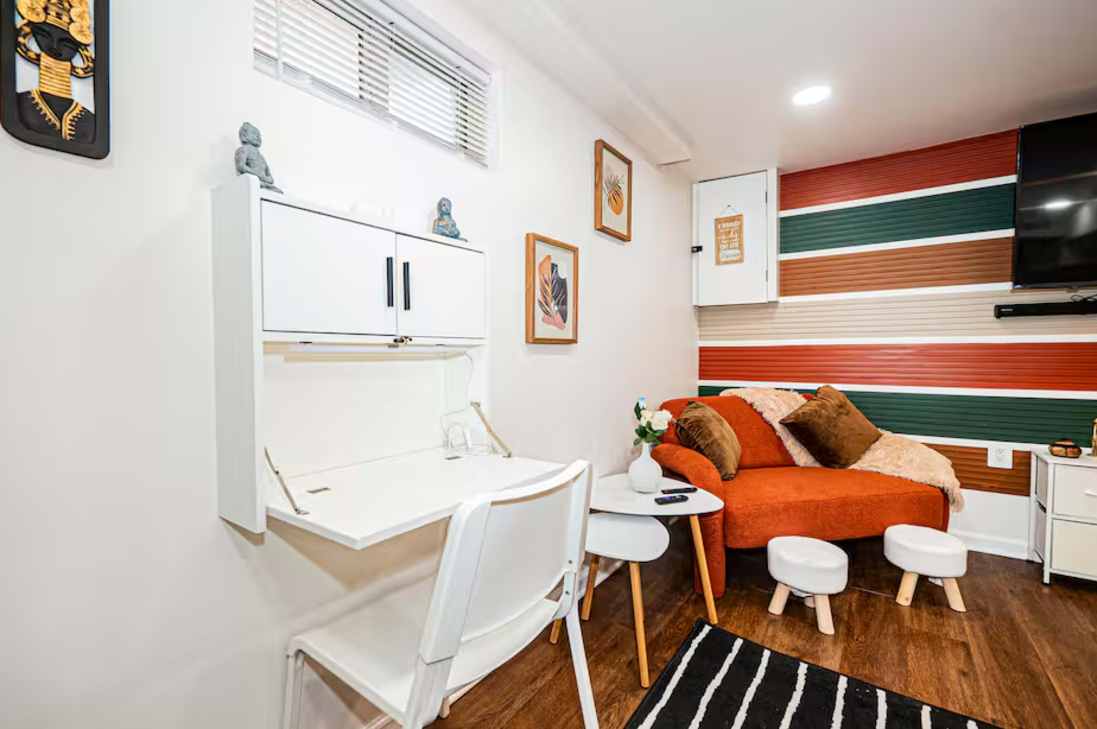 The image features a cozy room with a small desk and chair, an orange sofa, and colorful striped wall decor.