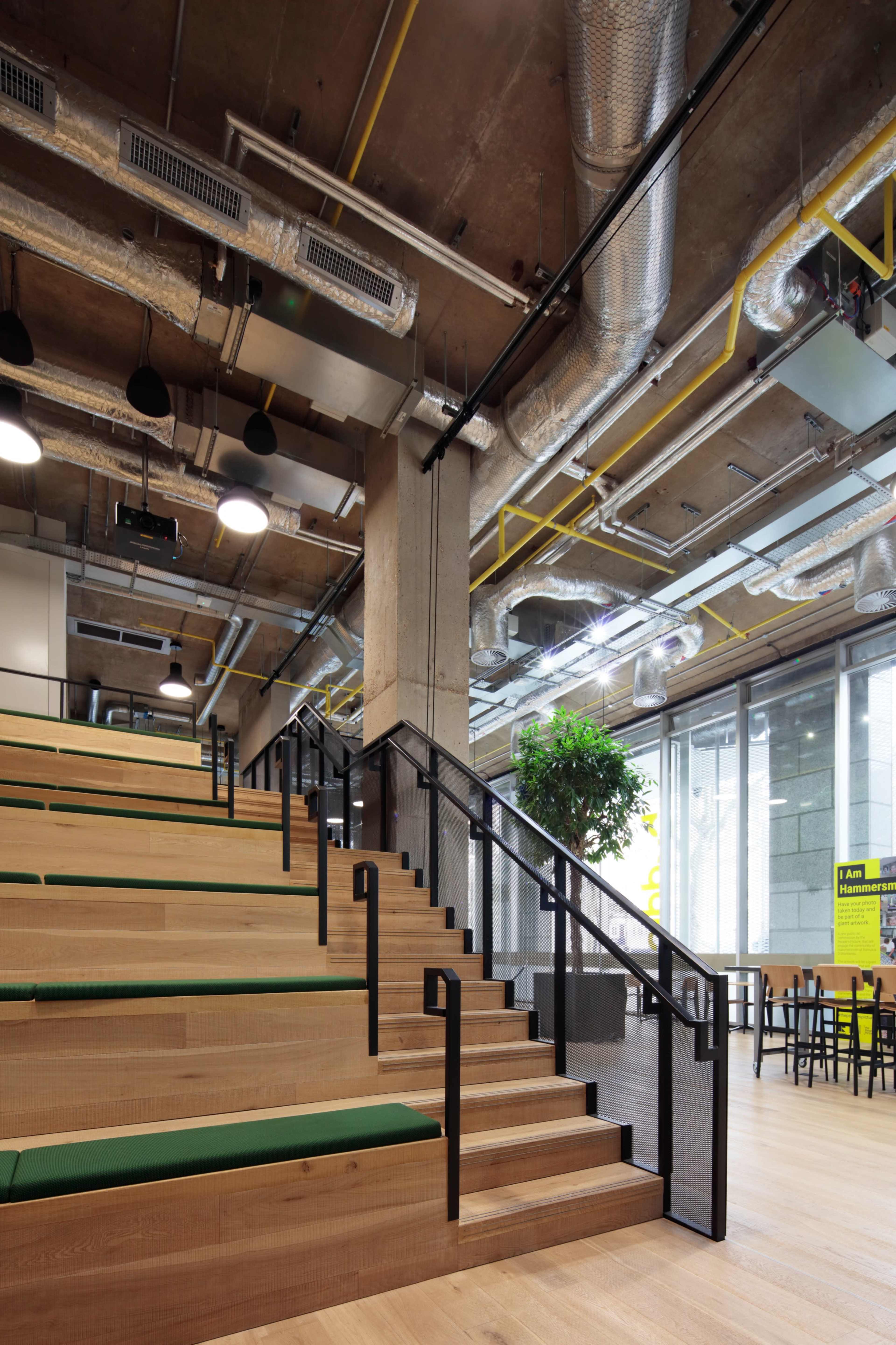A modern interior space featuring wooden tiered seating with green cushions, large windows, and exposed industrial ventilation systems.