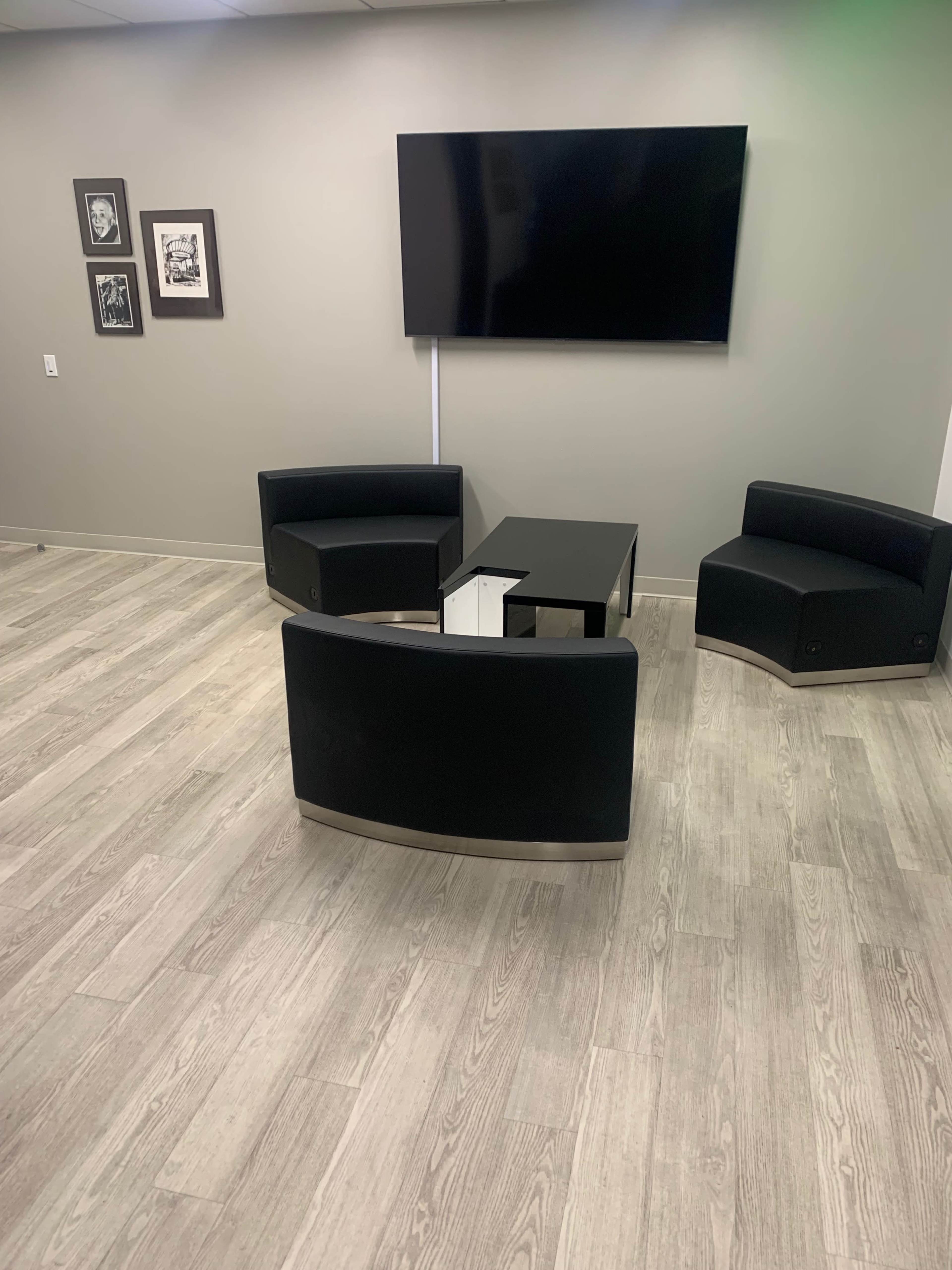 A modern seating area with two curved black chairs and a black coffee table, positioned on a light wooden floor under a wall-mounted television.