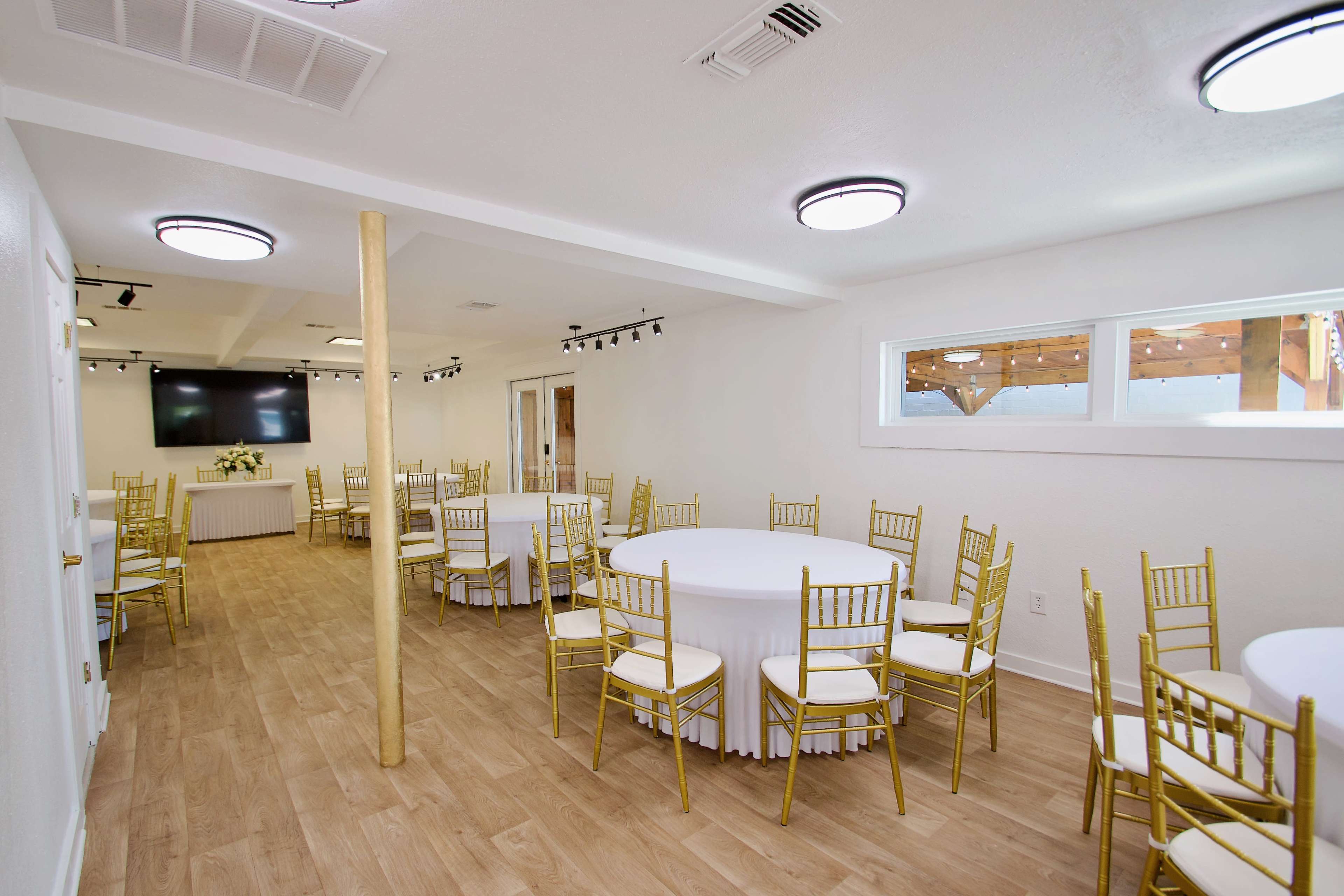 The image shows a spacious event room with round tables covered in white tablecloths and gold chairs arranged in an organized layout.