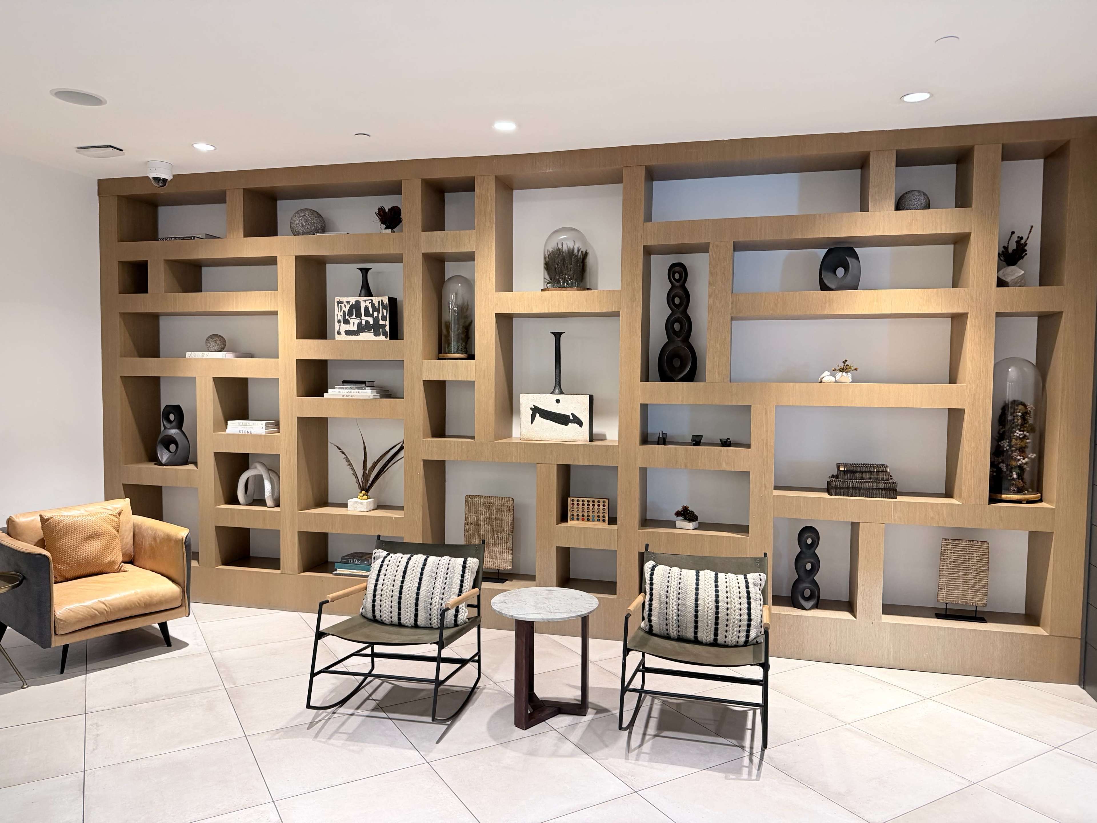 The image shows a modern interior with a wooden shelving unit displaying various decorative items, flanked by two lounge chairs and a small table on a tiled floor.