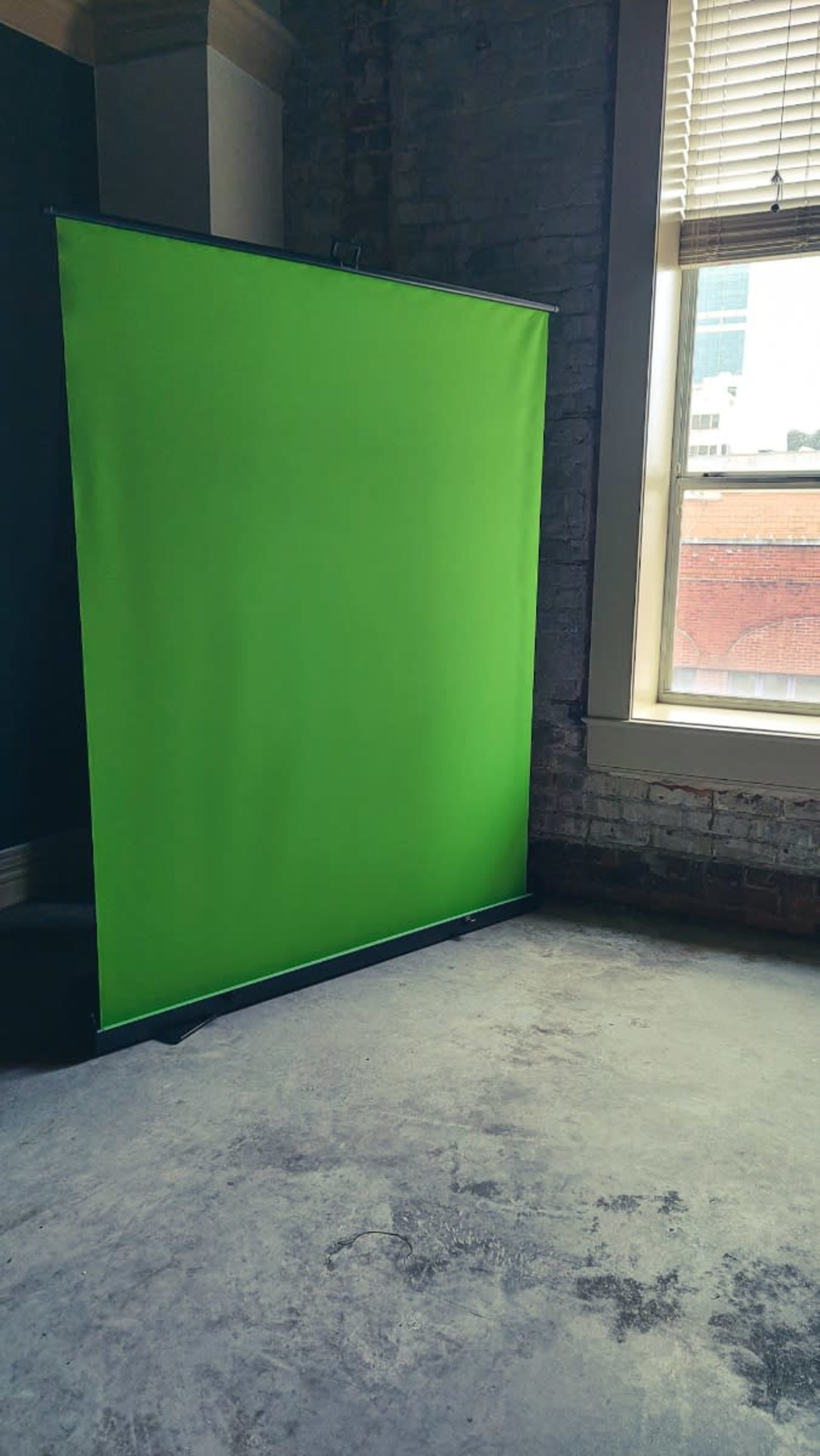 A green backdrop stands in a corner of a room with brick walls and a window letting in sunlight.