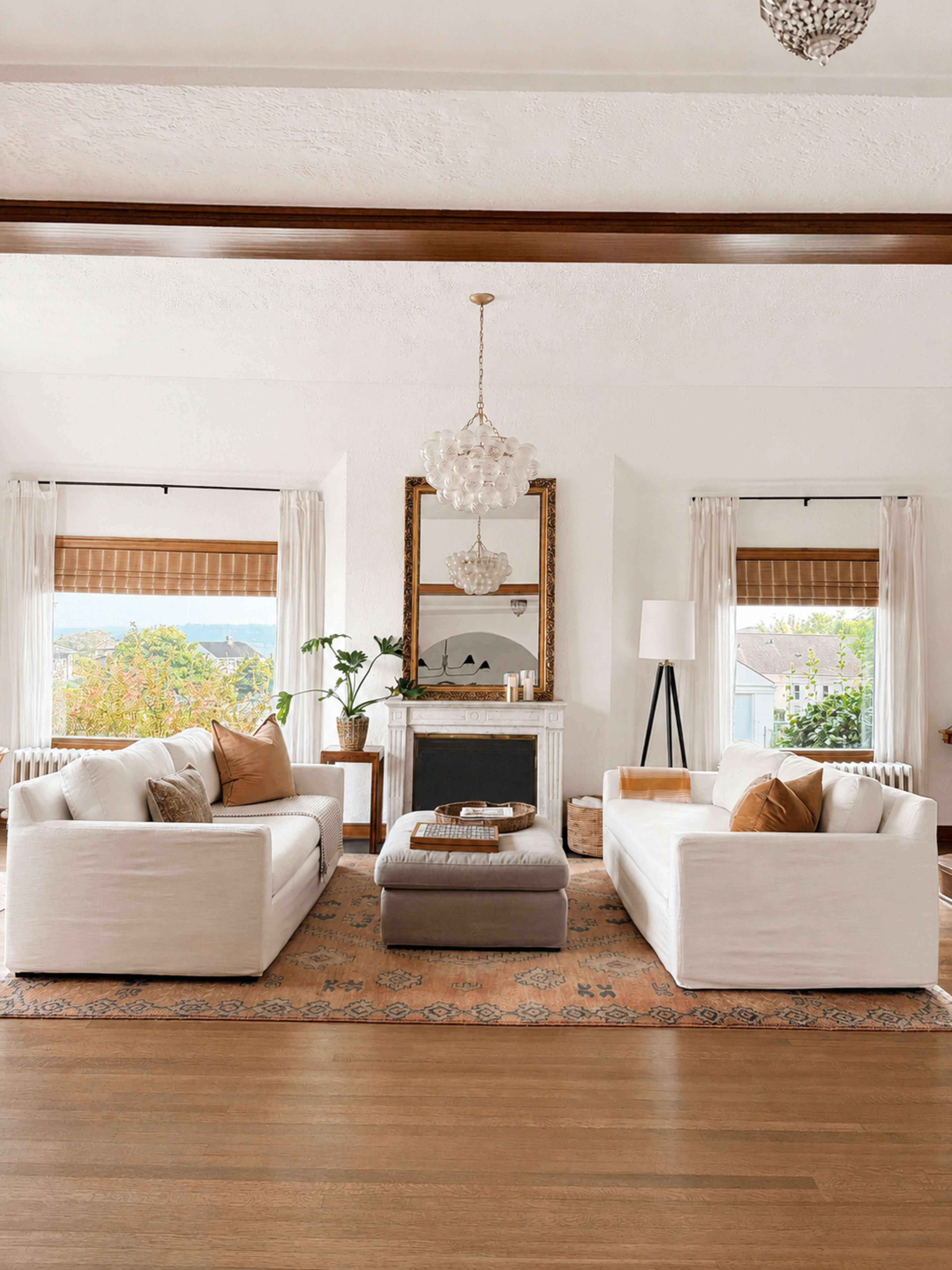 The living room features two white sofas facing each other, a central ottoman, a fireplace with a large mirror above it, and large windows dressed with shades, revealing an outdoor view.