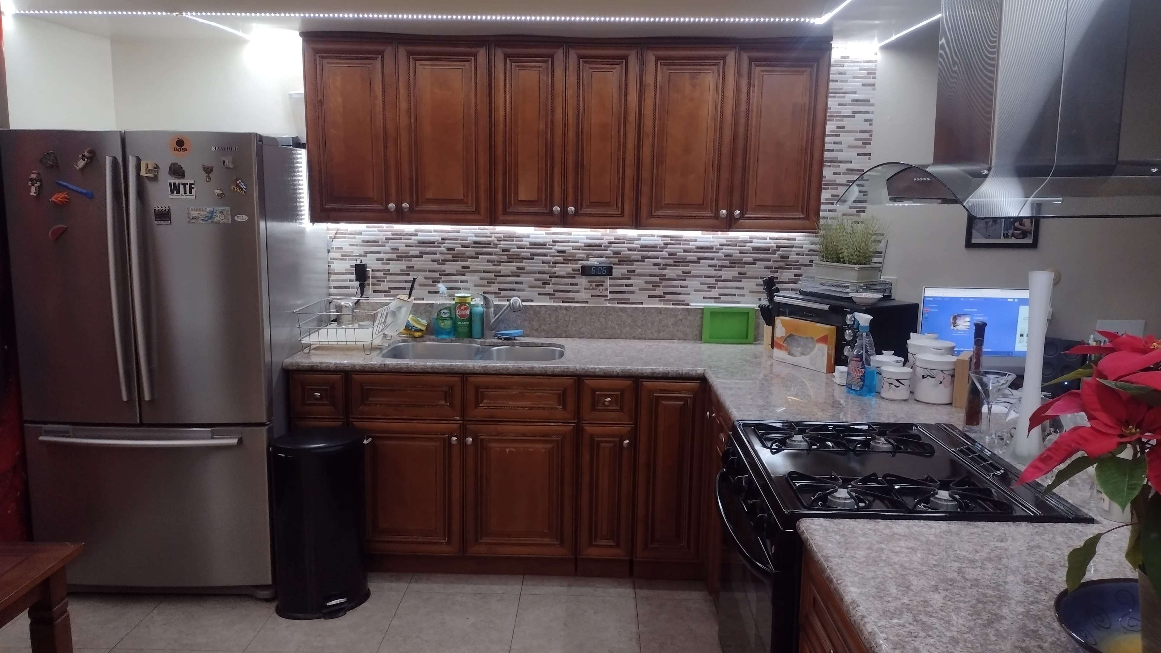 A kitchen with wooden cabinets, a stainless steel refrigerator, a sink with dish soap, and a gas stove with a granite countertop.