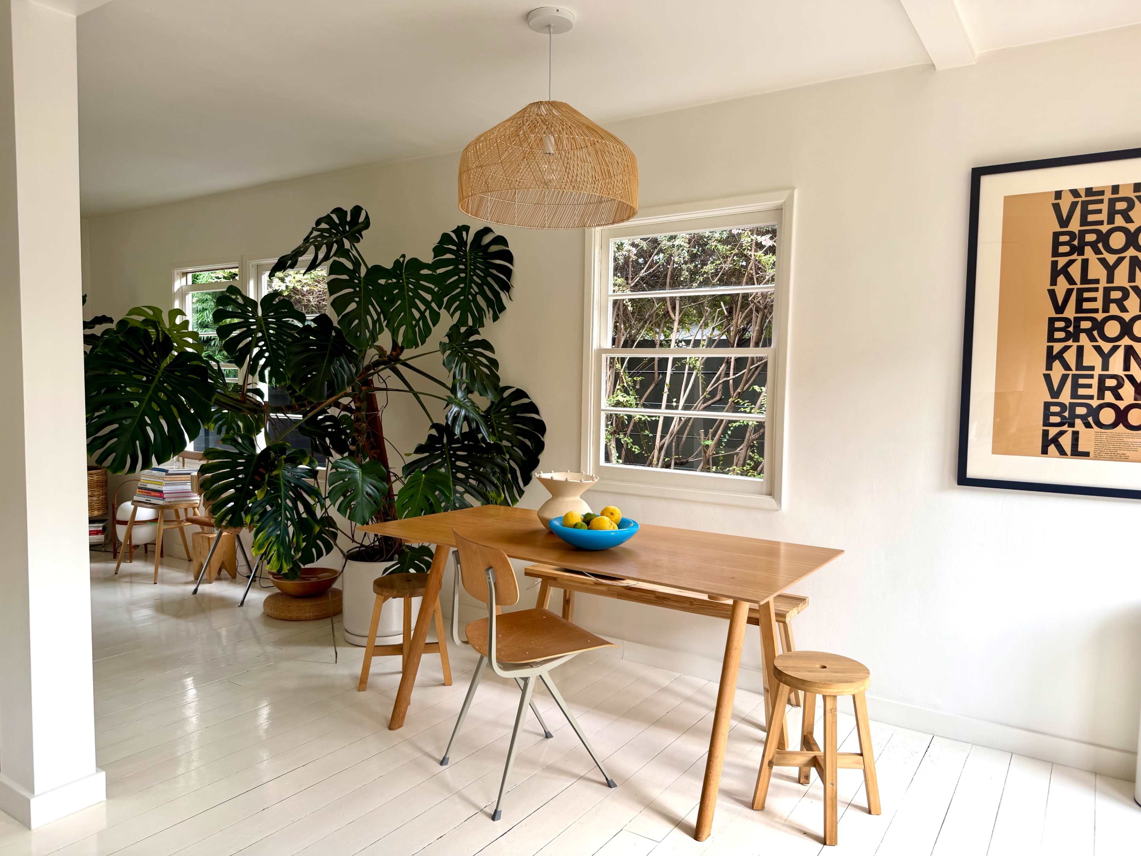 A wooden dining table with a blue bowl of fruit is set against a backdrop of large windows and a tall green plant in a well-lit room.