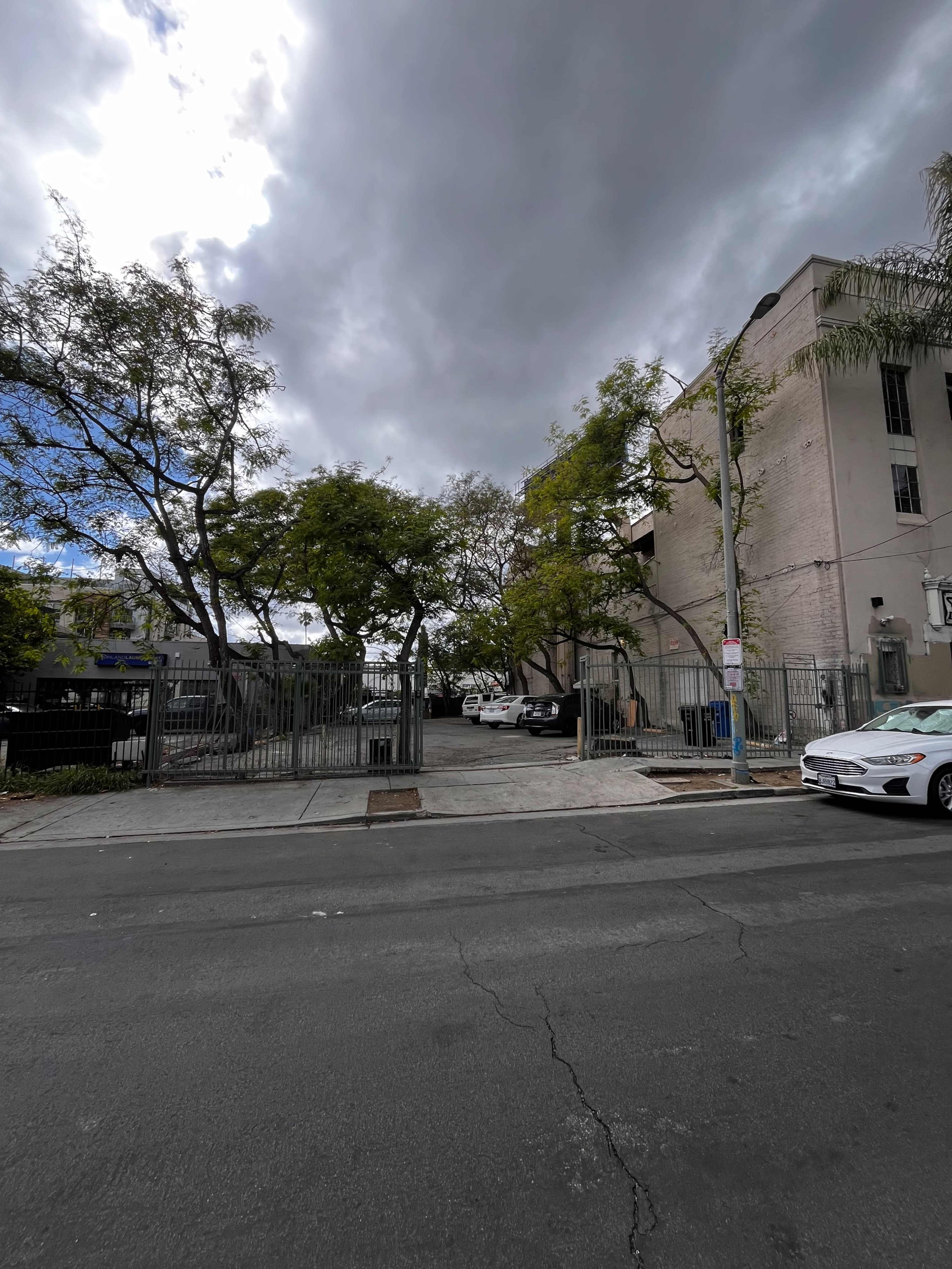 The image shows a street corner with a gated entrance to a parking area, flanked by trees and a multi-story building under a cloudy sky.