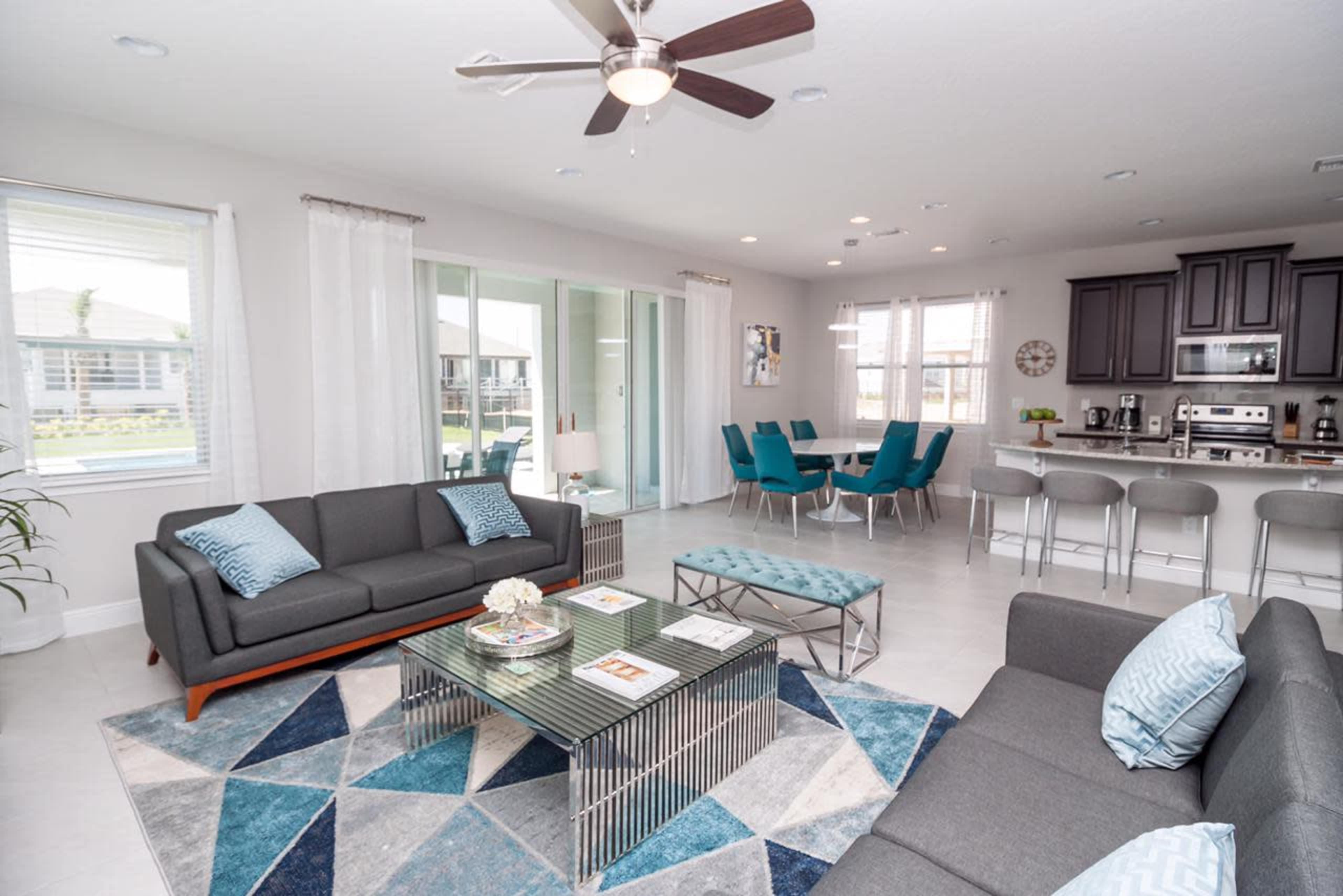 A modern living room with two sofas, a coffee table, a decorative area rug, and a view into a dining area with teal chairs and a kitchen.