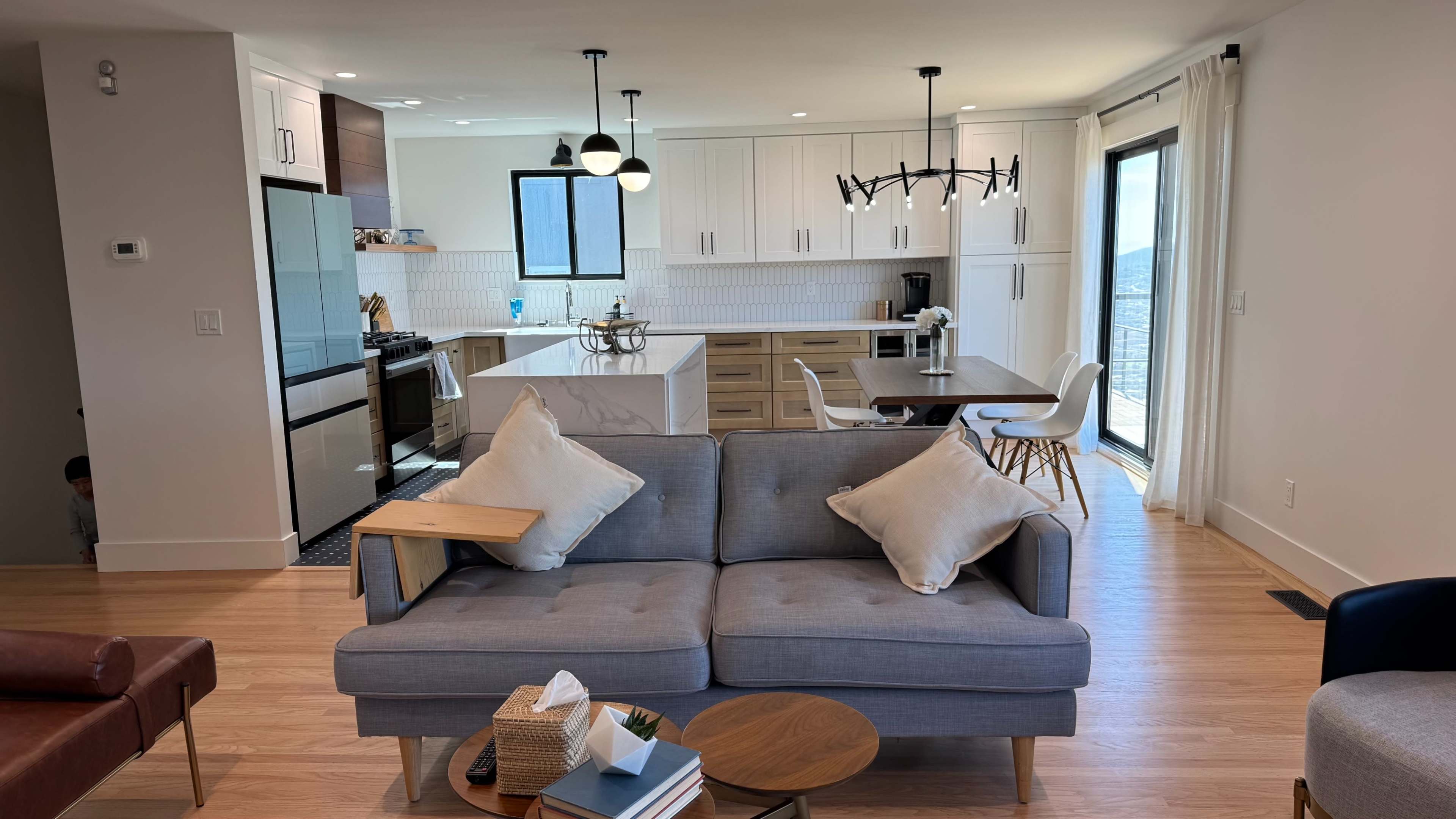 The image shows a modern, open-concept living space with a gray sofa, wooden dining table, and a kitchen with white cabinetry and stainless steel appliances.