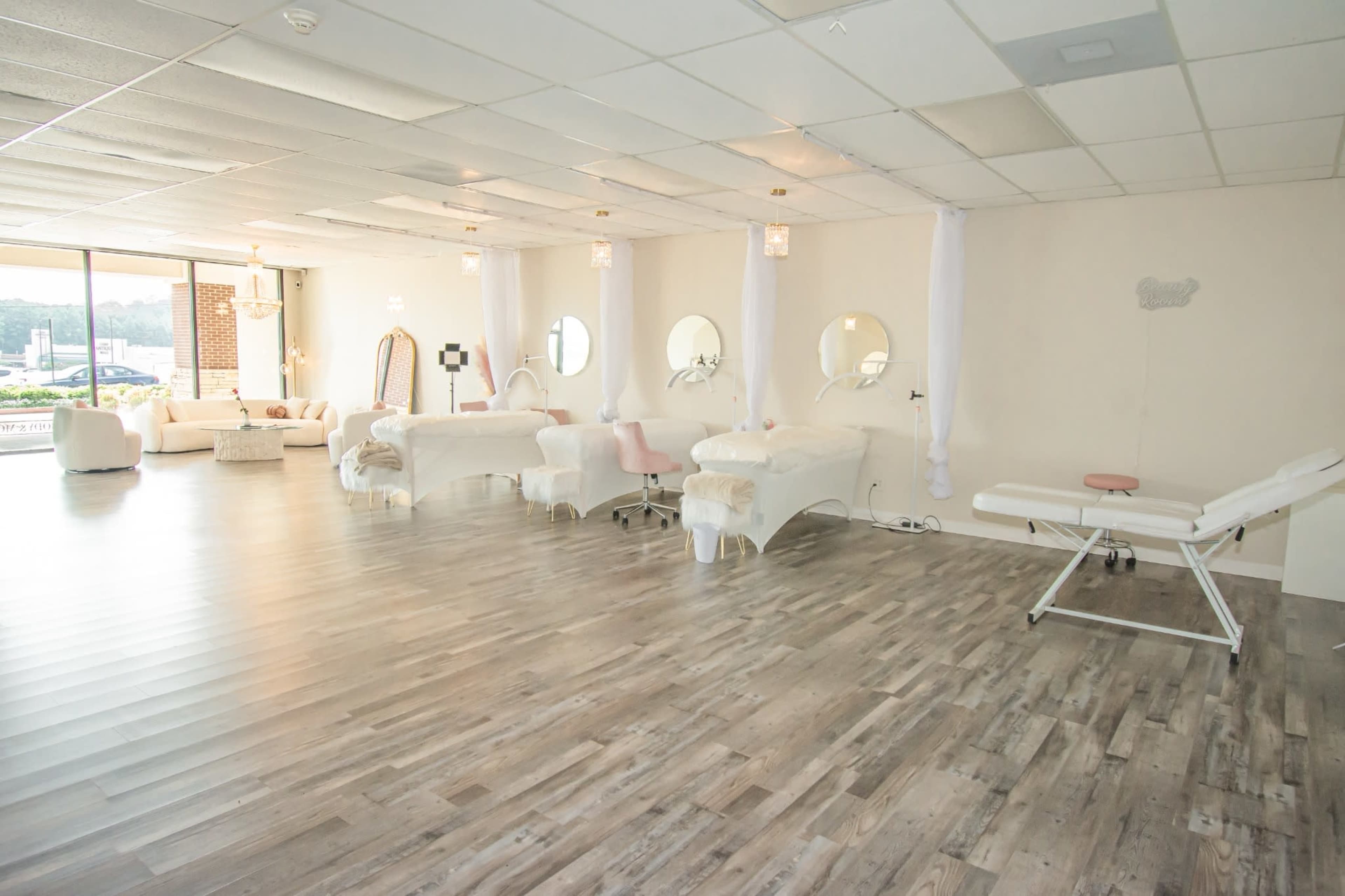 The image shows a spacious beauty treatment room with multiple treatment beds, large windows, and a cozy seating area.