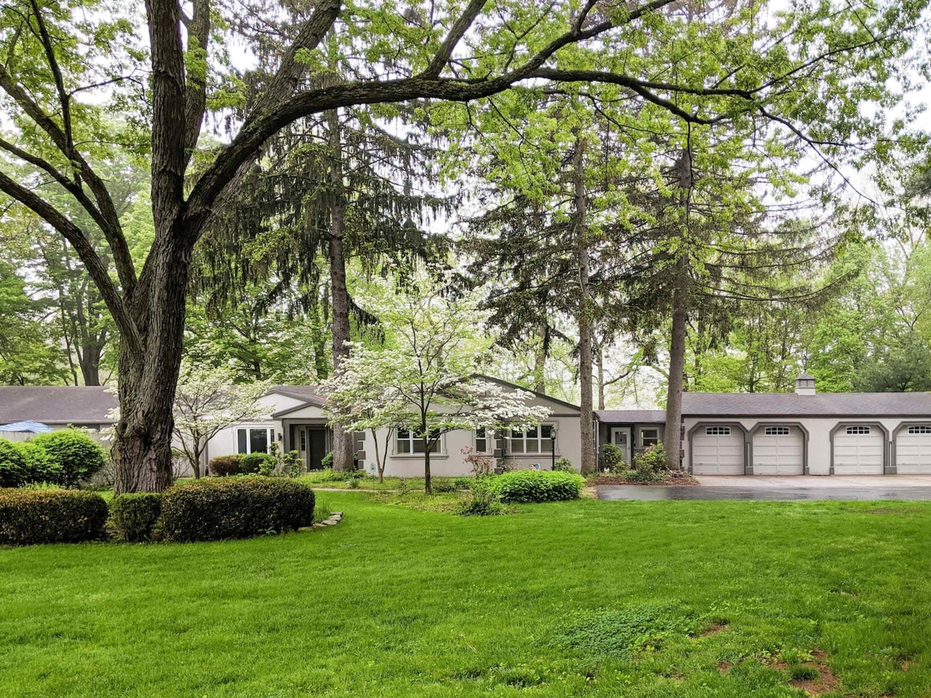 A single-story house is surrounded by lush greenery and trees, with a driveway leading to a three-car garage.