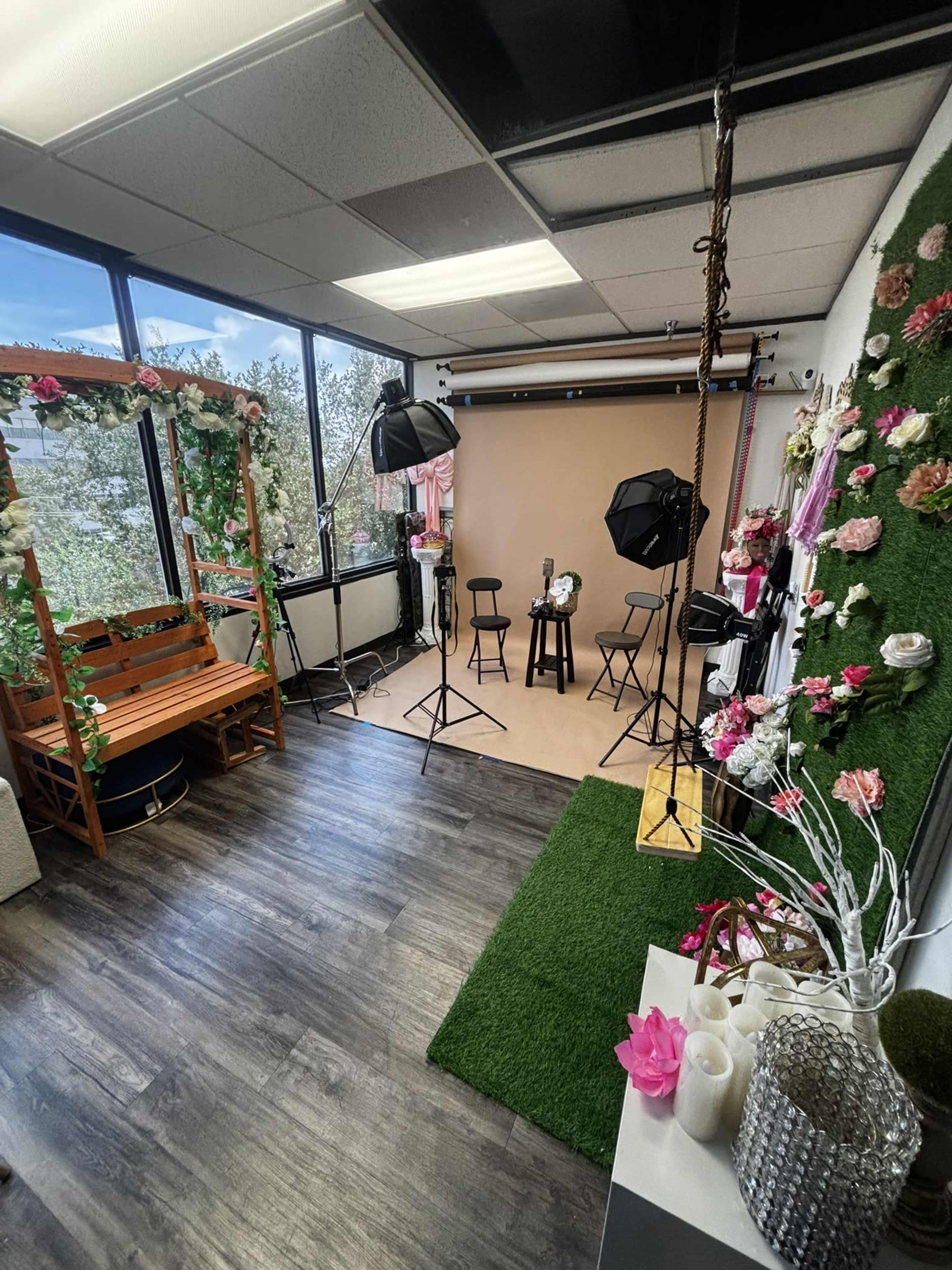 The image shows a photography studio featuring a backdrop, various lighting equipment, and decor elements such as artificial flowers and a wooden bench.