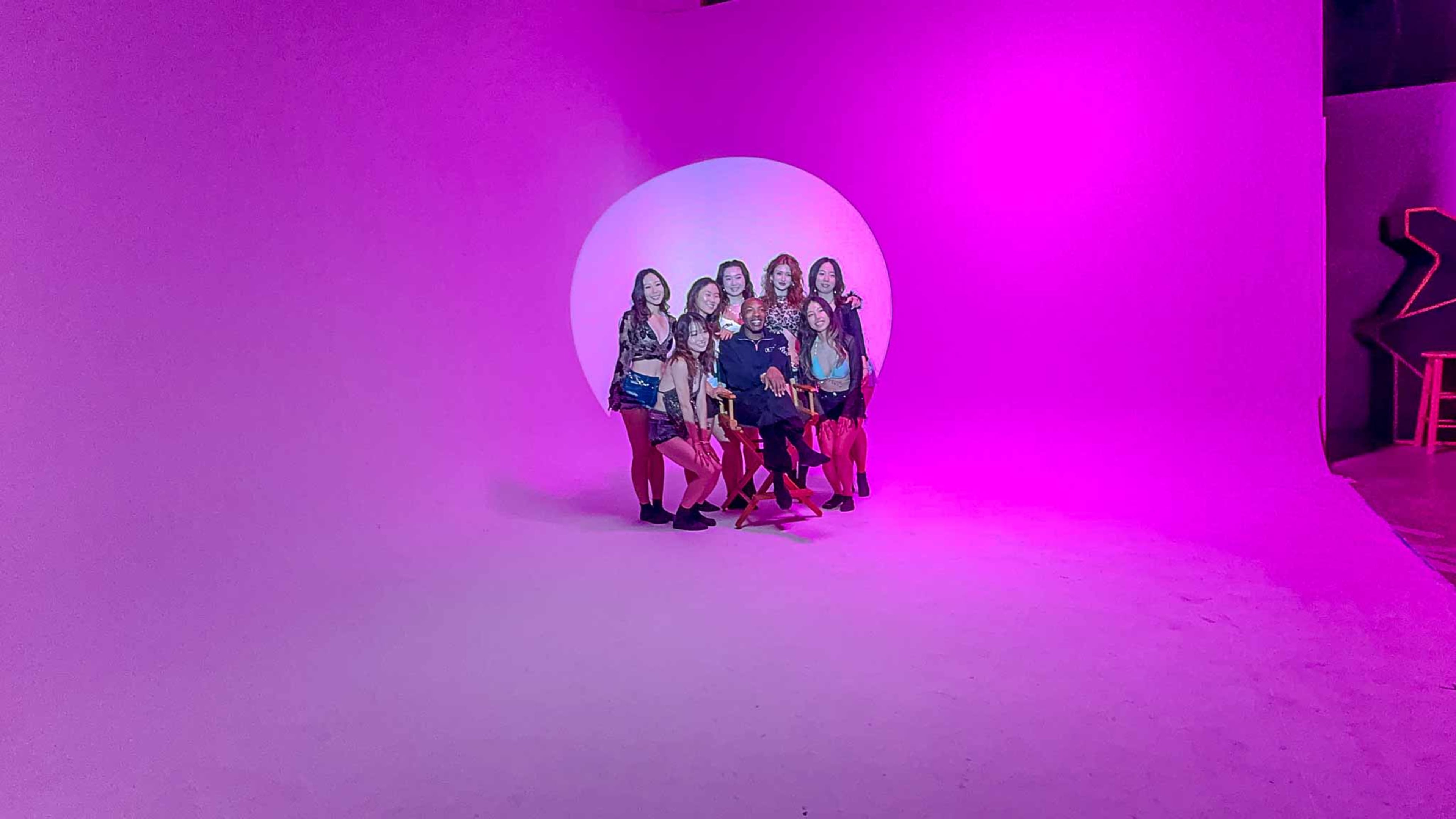 A group of young women poses together in front of a large, illuminated pink backdrop.