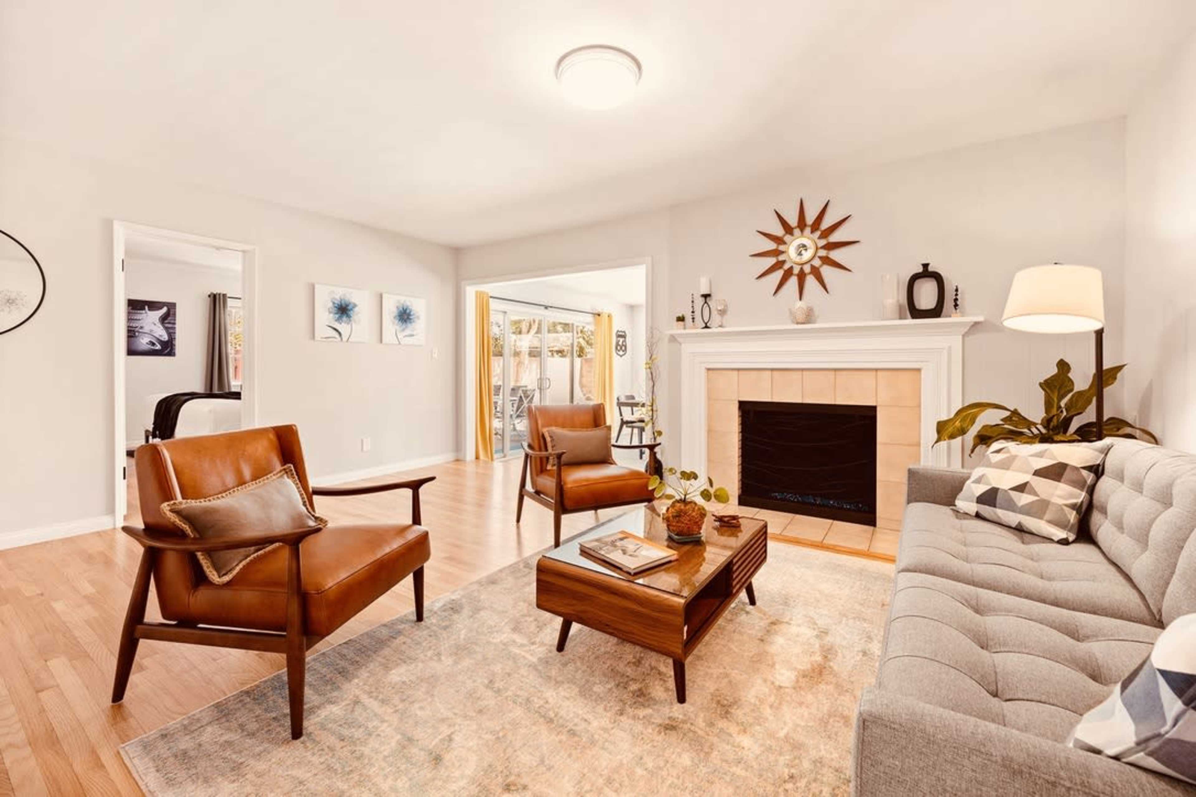 A modern living room features a gray sofa, brown leather chairs, a wooden coffee table, a stone fireplace, and a decorative sunburst wall clock.