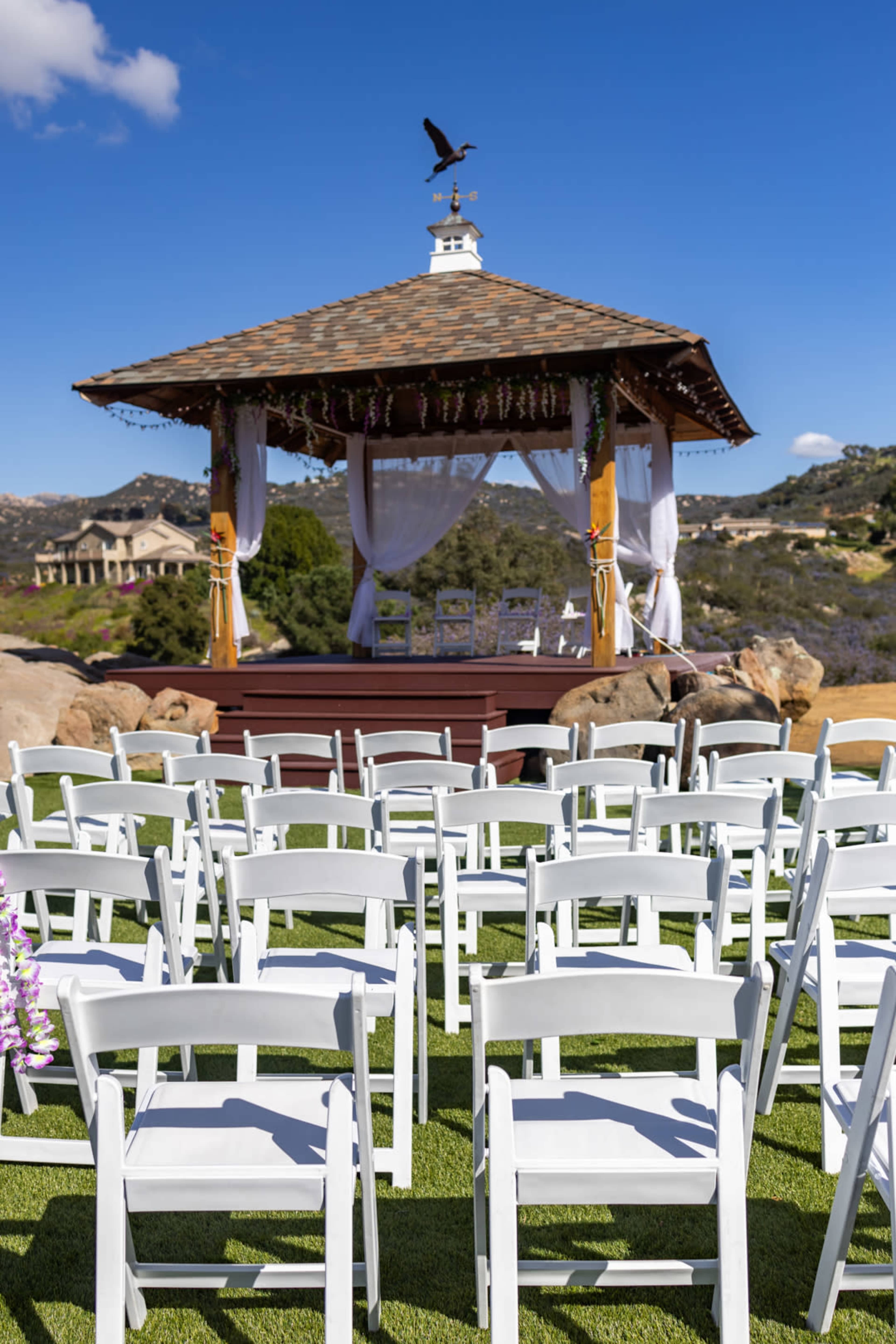 A gazebo with decorative drapes and floral accents stands on a grassy area, surrounded by rows of white chairs and picturesque hills in the background.