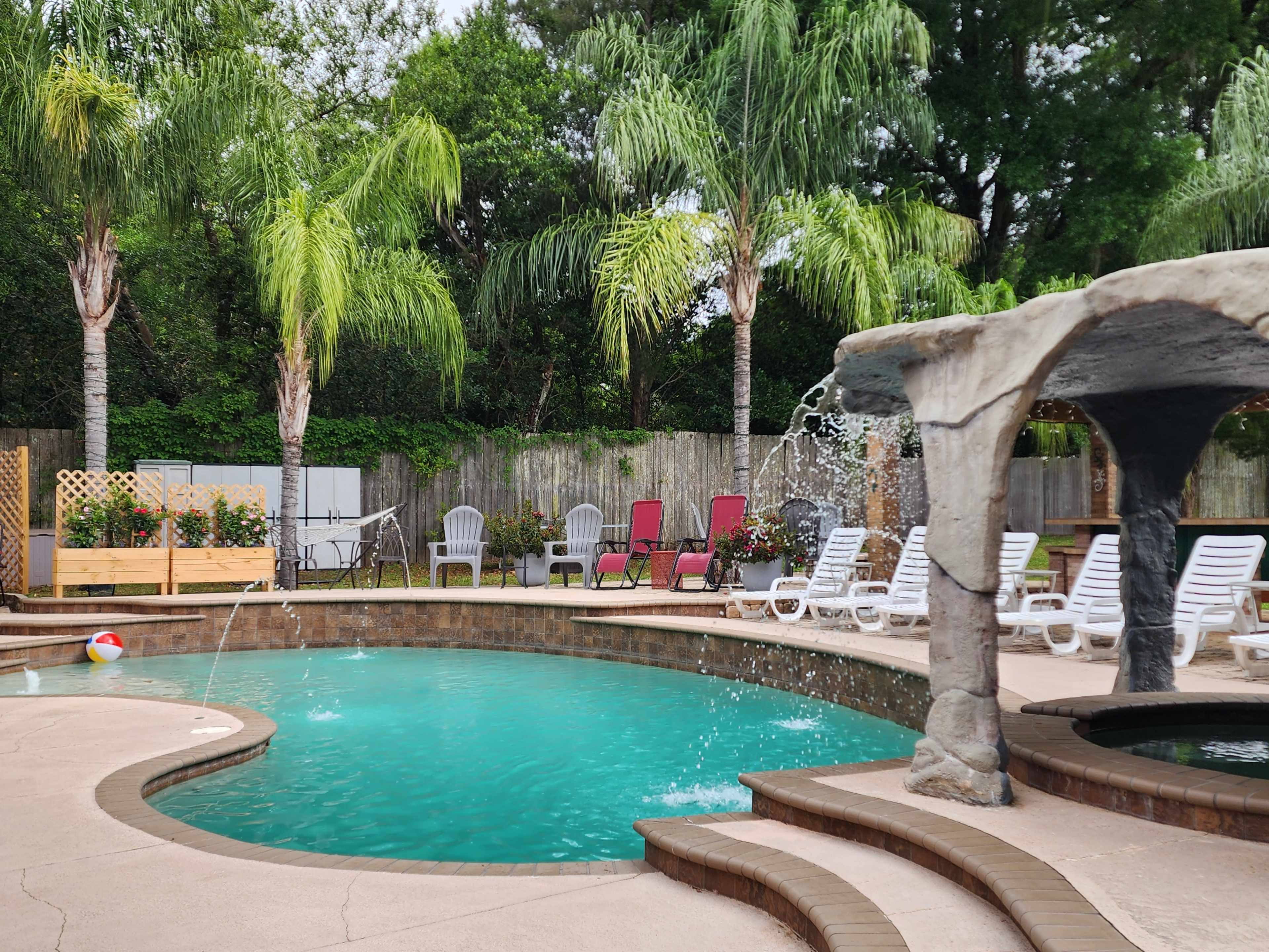 The image shows a swimming pool surrounded by lounge chairs and palm trees, with a rock fountain and a beach ball floating in the water.