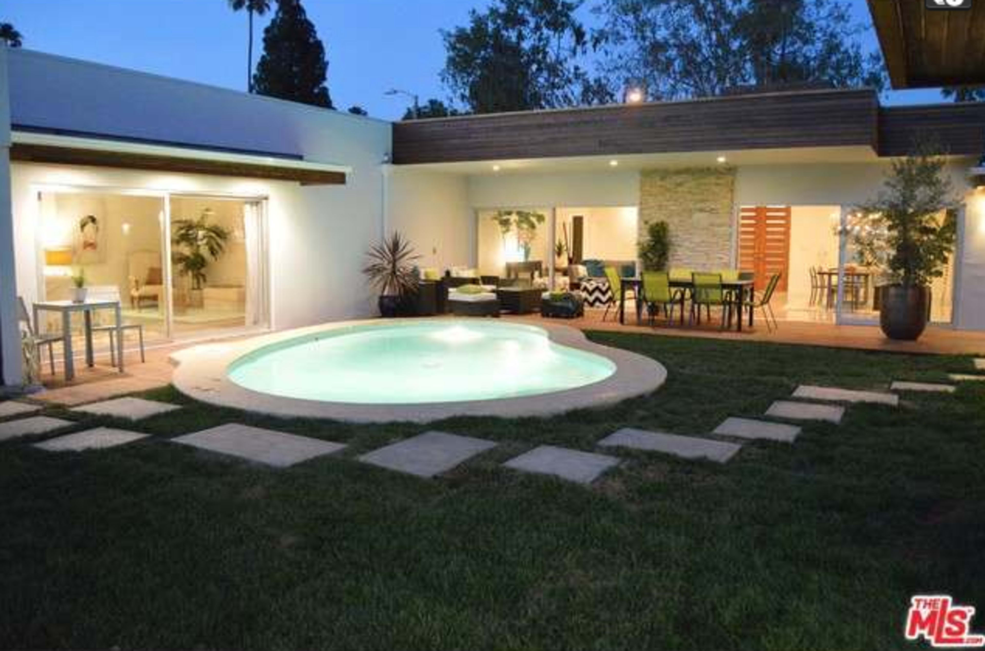 The image shows a modern home with a circular pool surrounded by grass and stone pathways, featuring sliding glass doors that open to an outdoor seating area.