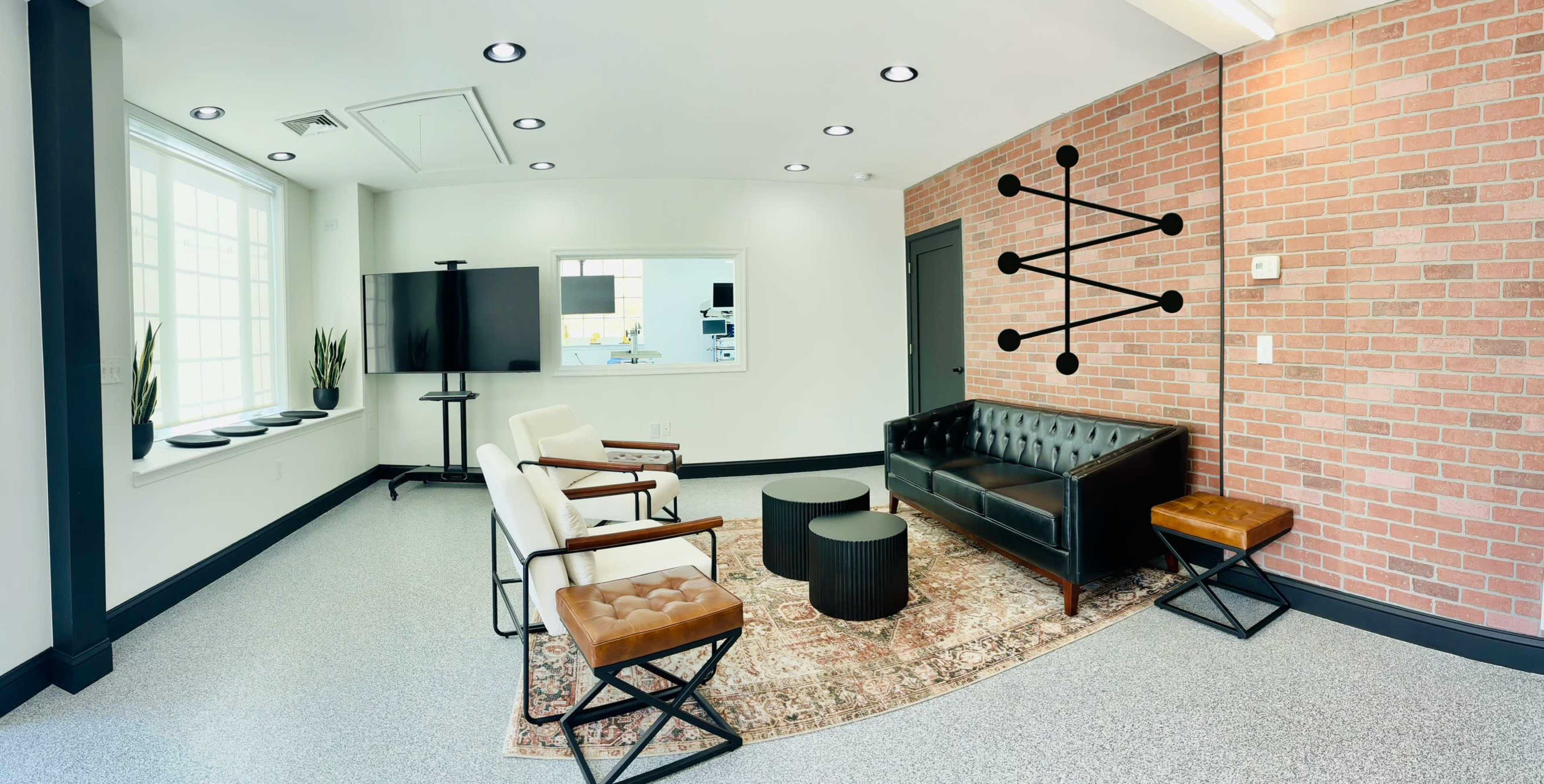 The image shows a modern, minimalist lounge area featuring a black leather sofa, two armchairs, a round coffee table, and a patterned rug, along with a television mounted on the wall.