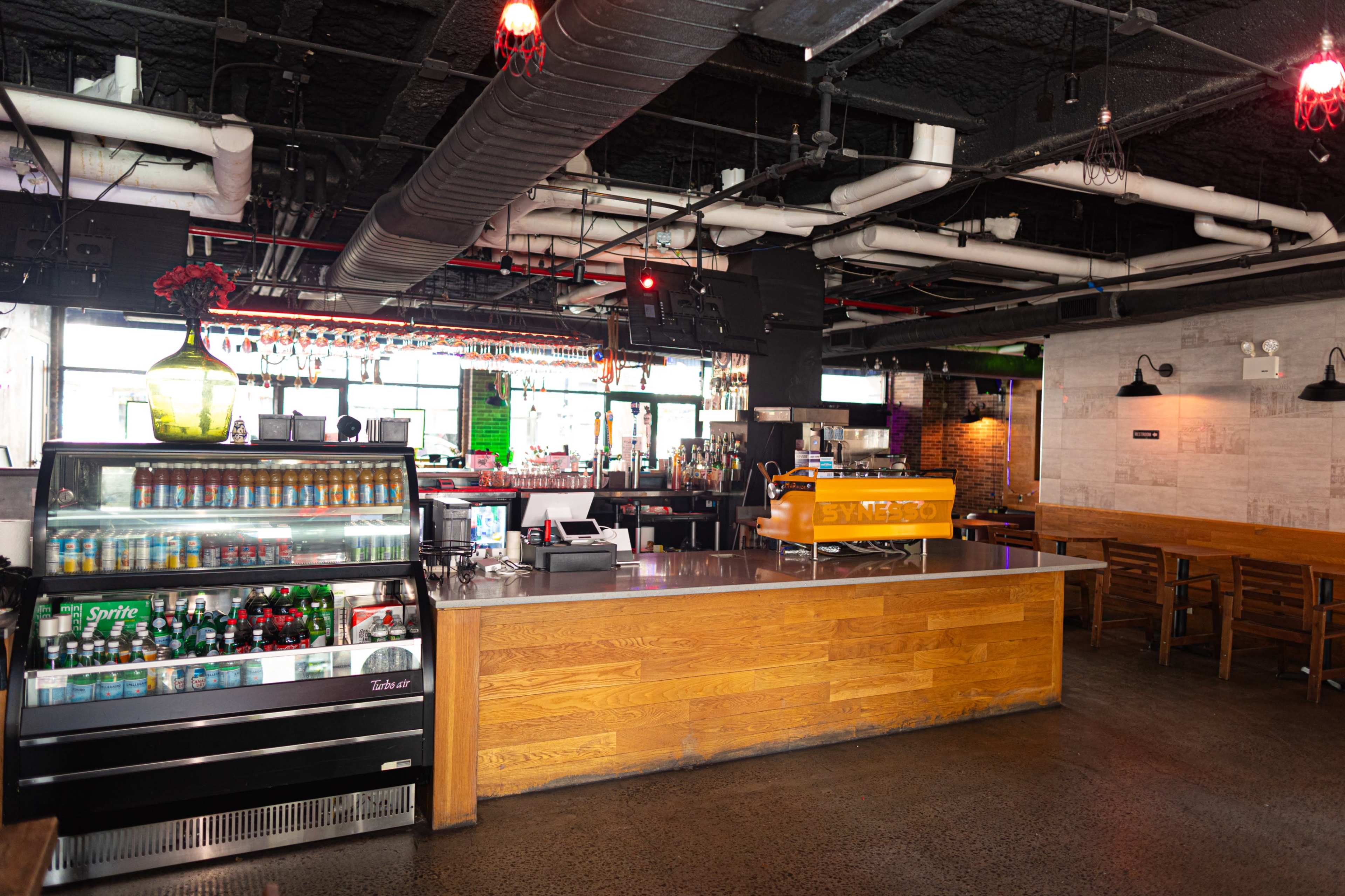 The image shows a modern restaurant interior featuring a bar area with a variety of beverage refrigerators and wooden seating arrangements.