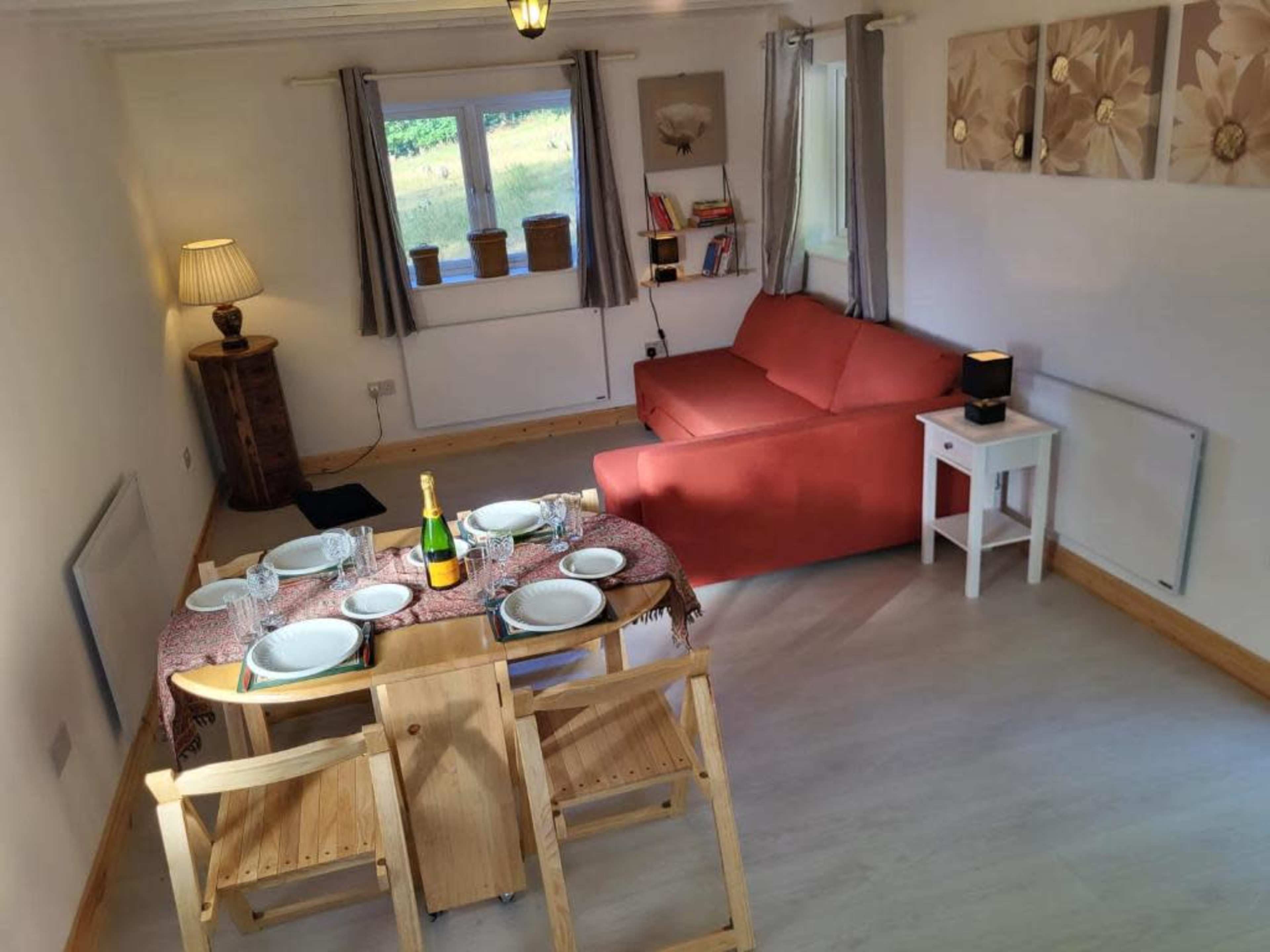 A cozy living space with a red couch, a dining table set for four, and a lamp in the corner, featuring light-colored walls and a view of greenery through the window.