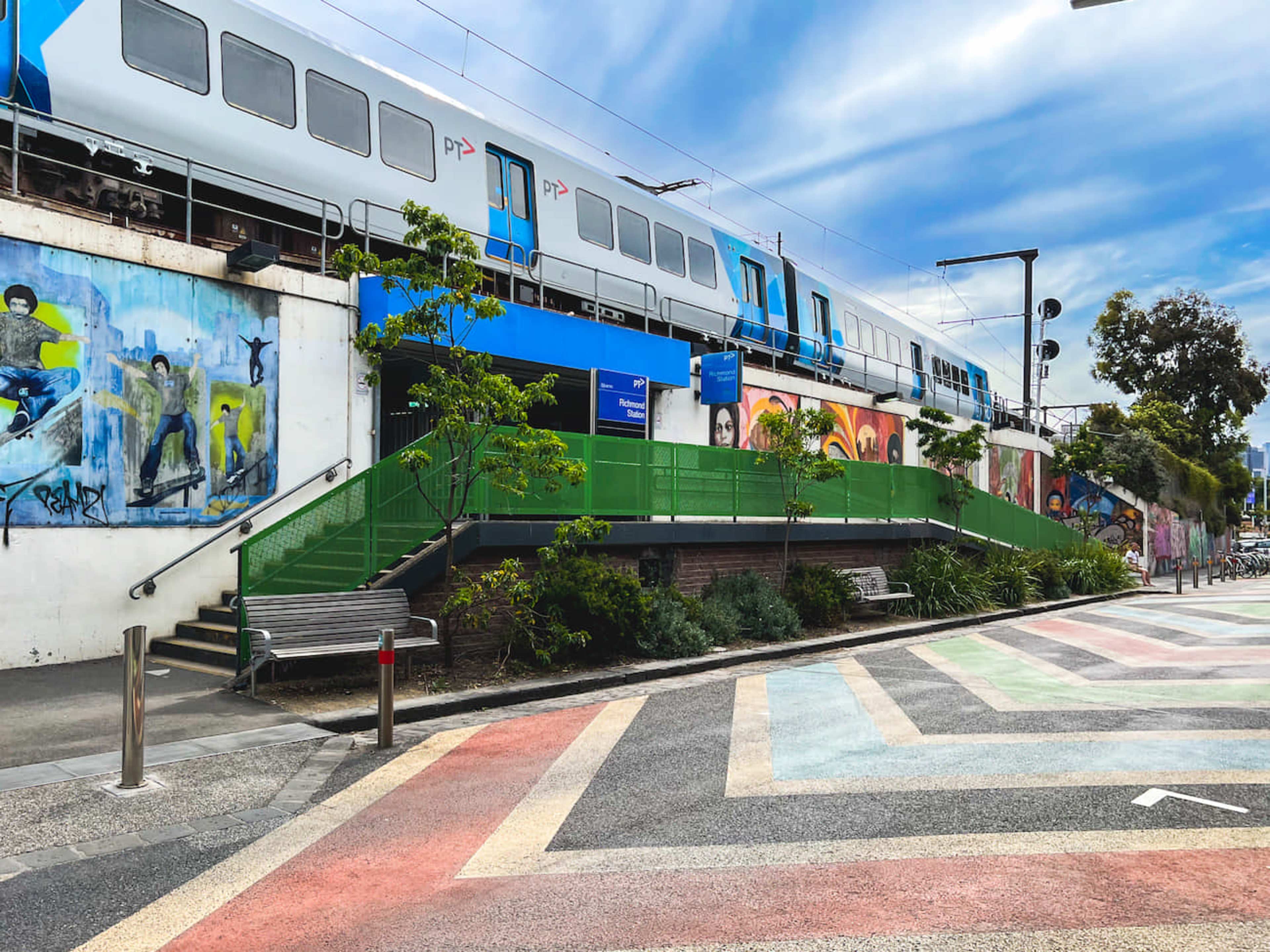 A train passes above a pedestrian area adorned with colorful street art and patterned pavement.