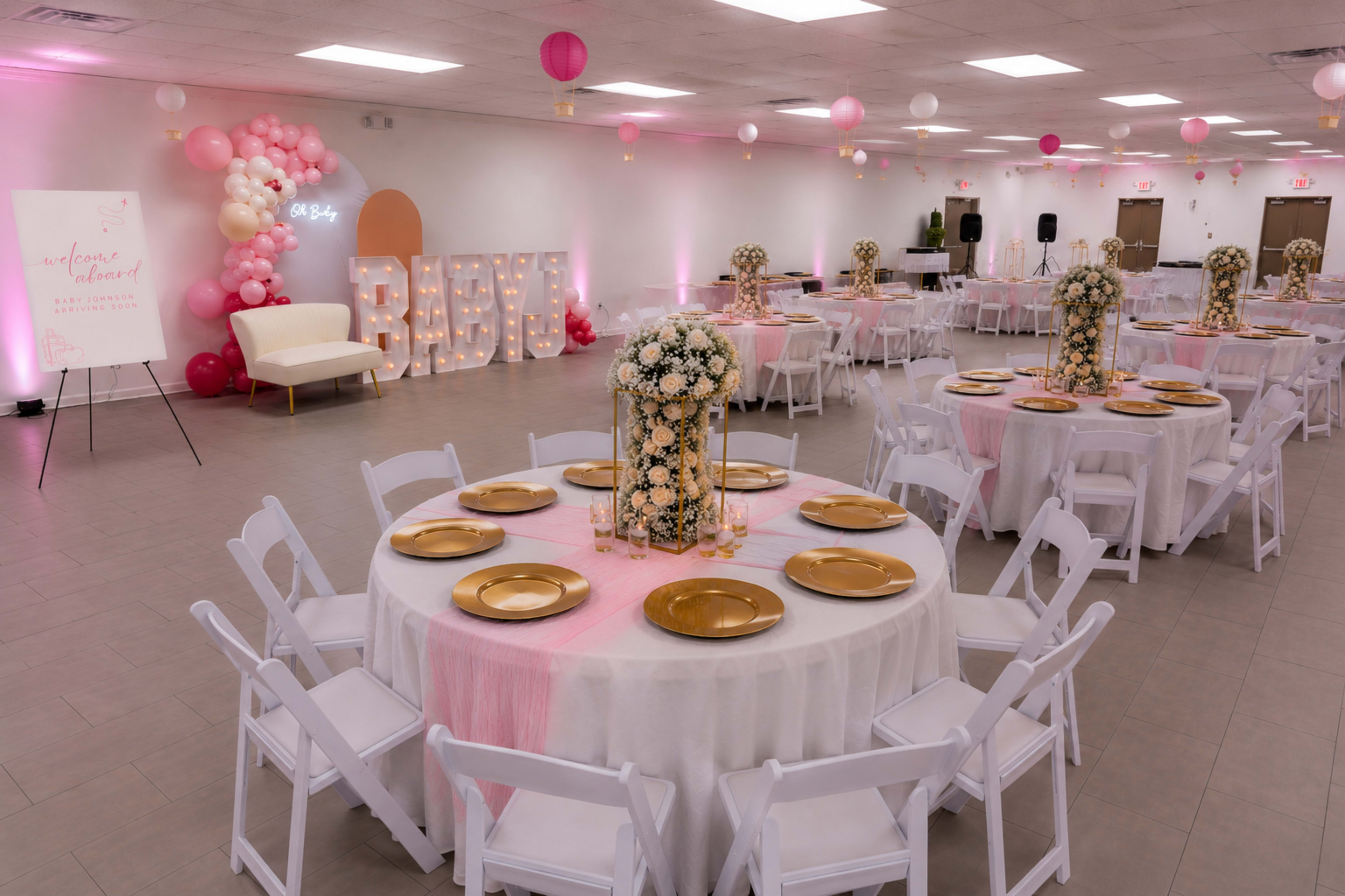 The image shows a decorated event space featuring round tables with white tablecloths and gold plates, surrounded by pink and gold decor, large light-up letters spelling "BABY," and floral centerpieces.