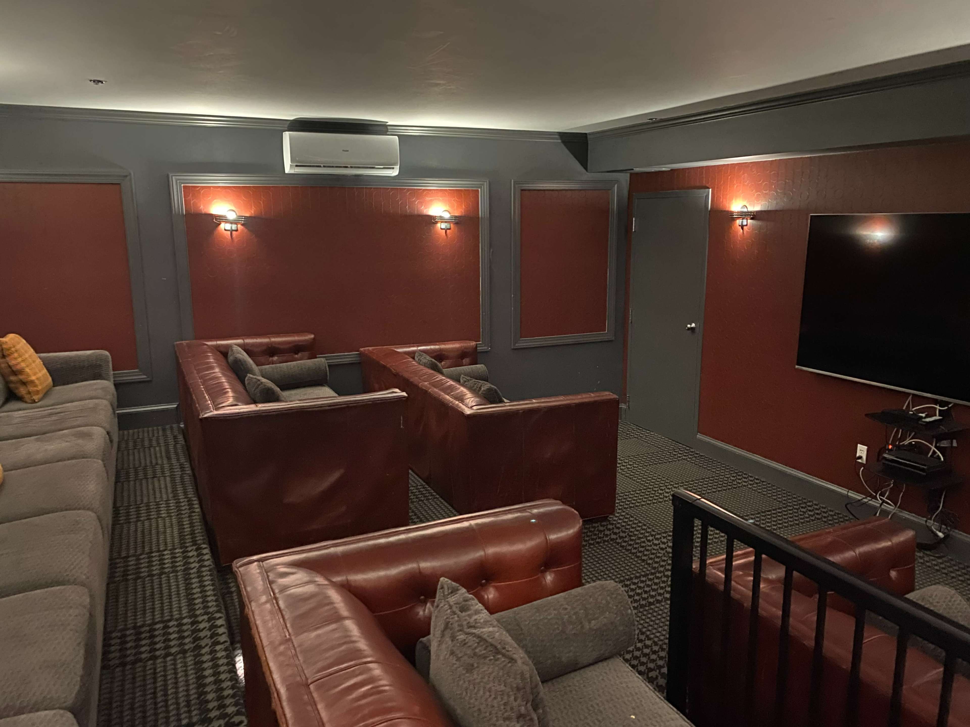 A theater-like room featuring several leather couches arranged around a large wall-mounted television, with dark red walls and dim lighting.